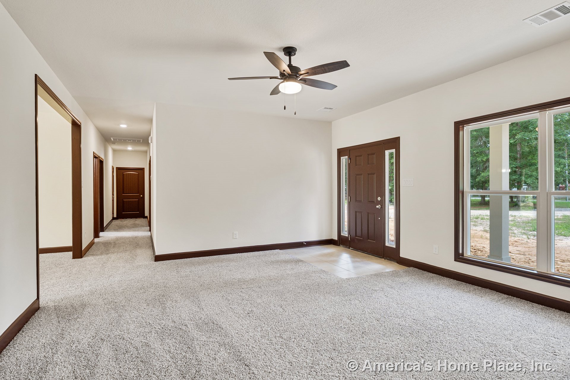Carpeted living room with ceiling fan and light fixture, brown trim and doors, large windows with brown frames, tile entryway featuring a brown front door with sidelights, and
