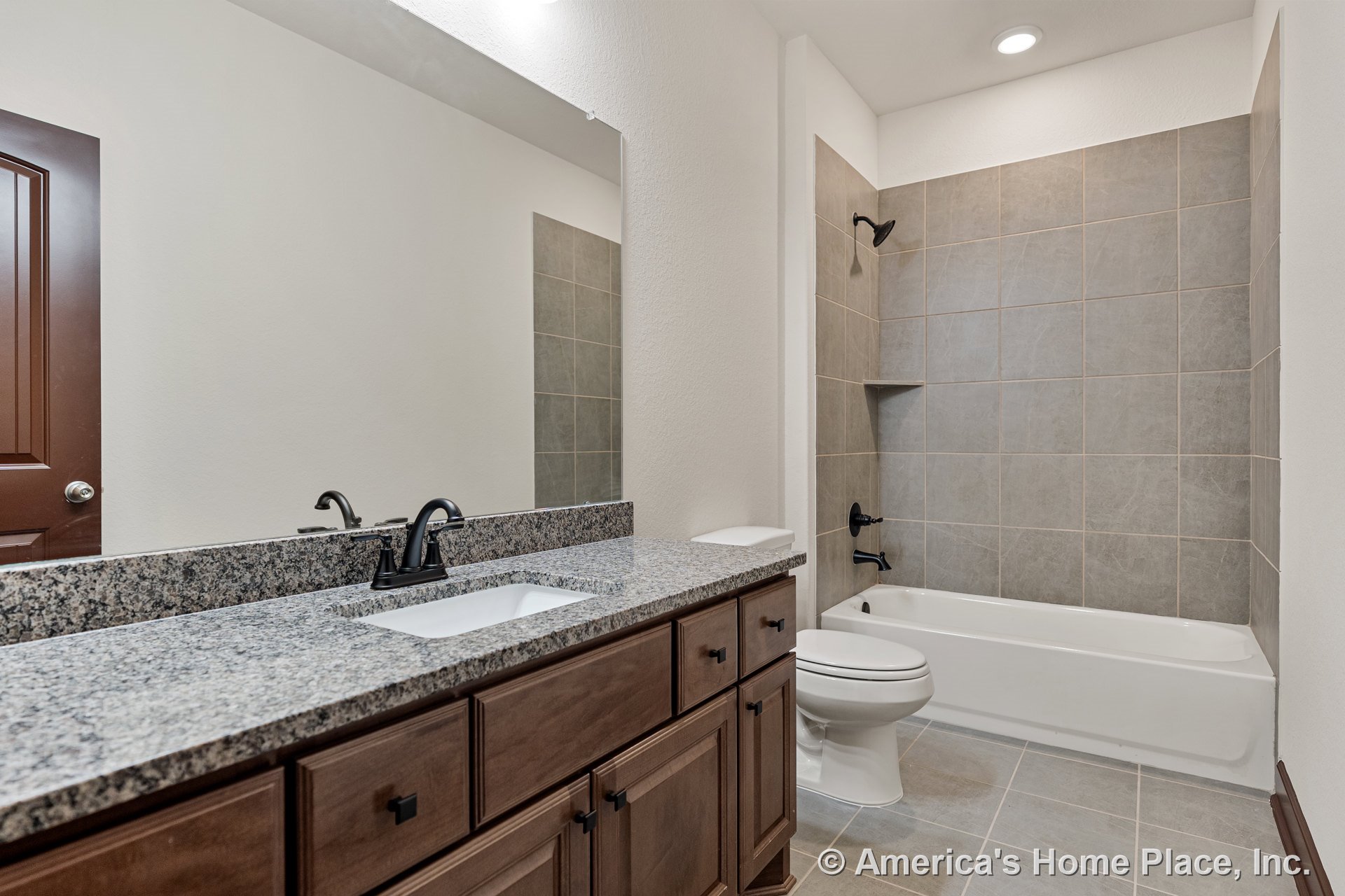 Granite countertop vanity with dark wood cabinetry, large wall mirror, single sink with modern dark faucet, tiled shower surround featuring built-in niche, white bathtub, ceramic