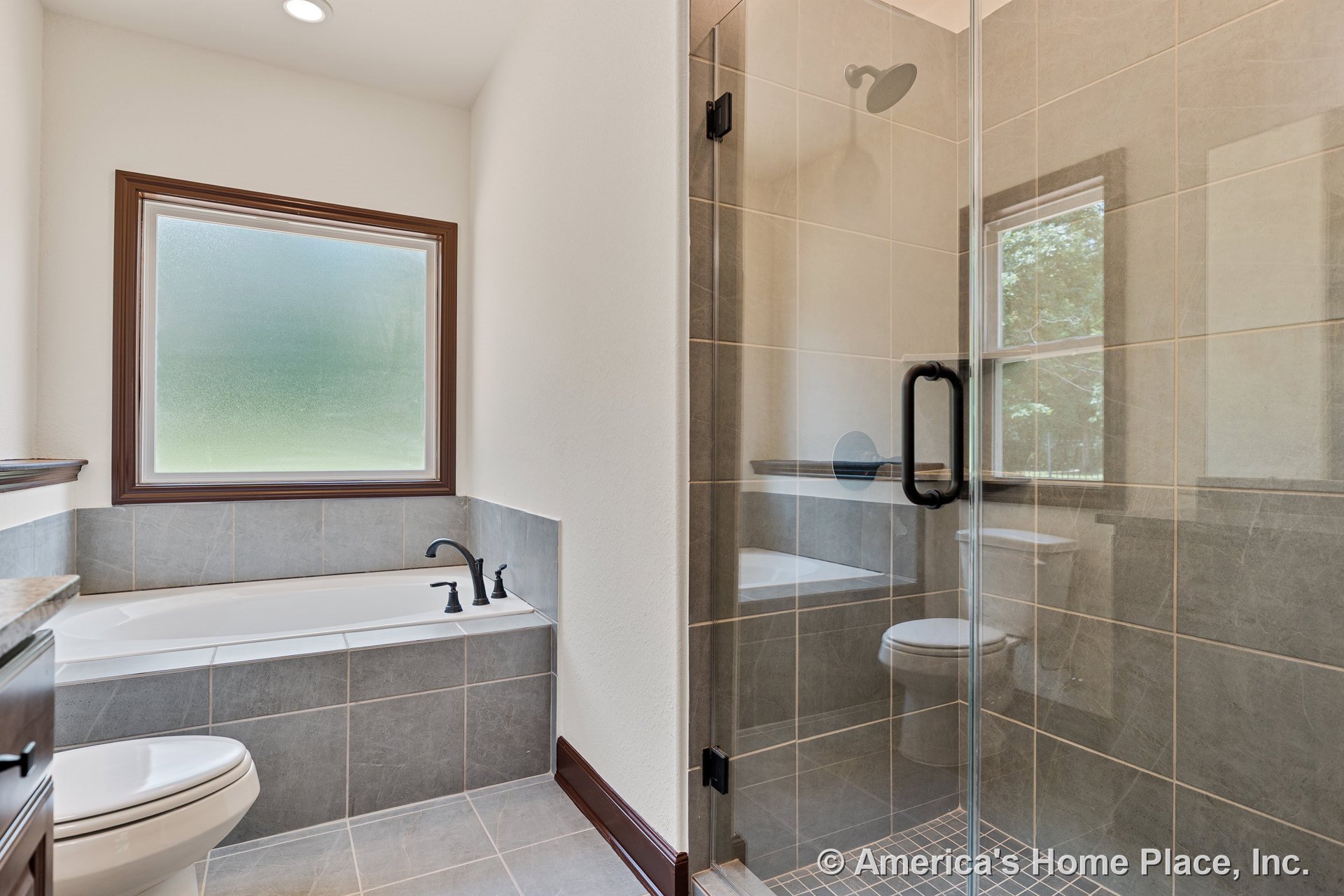 Glass-enclosed tiled shower beside a built-in bathtub under a frosted window, gray tile flooring, dark wood trim, and recessed ceiling lighting.