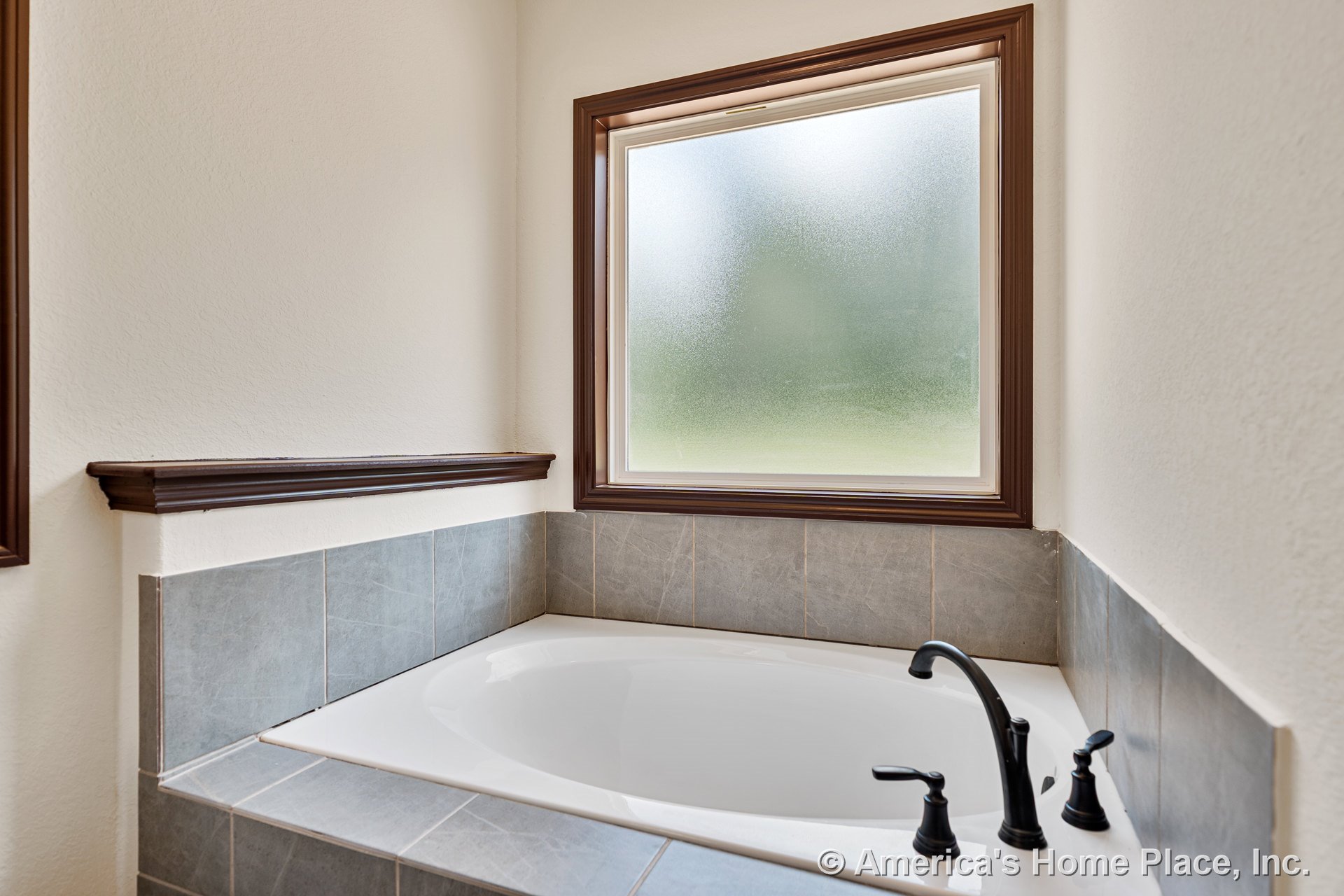 Oval soaking tub with gray tile surround and dark bronze faucet, set in a bathroom corner beneath a frosted window with wood trim.