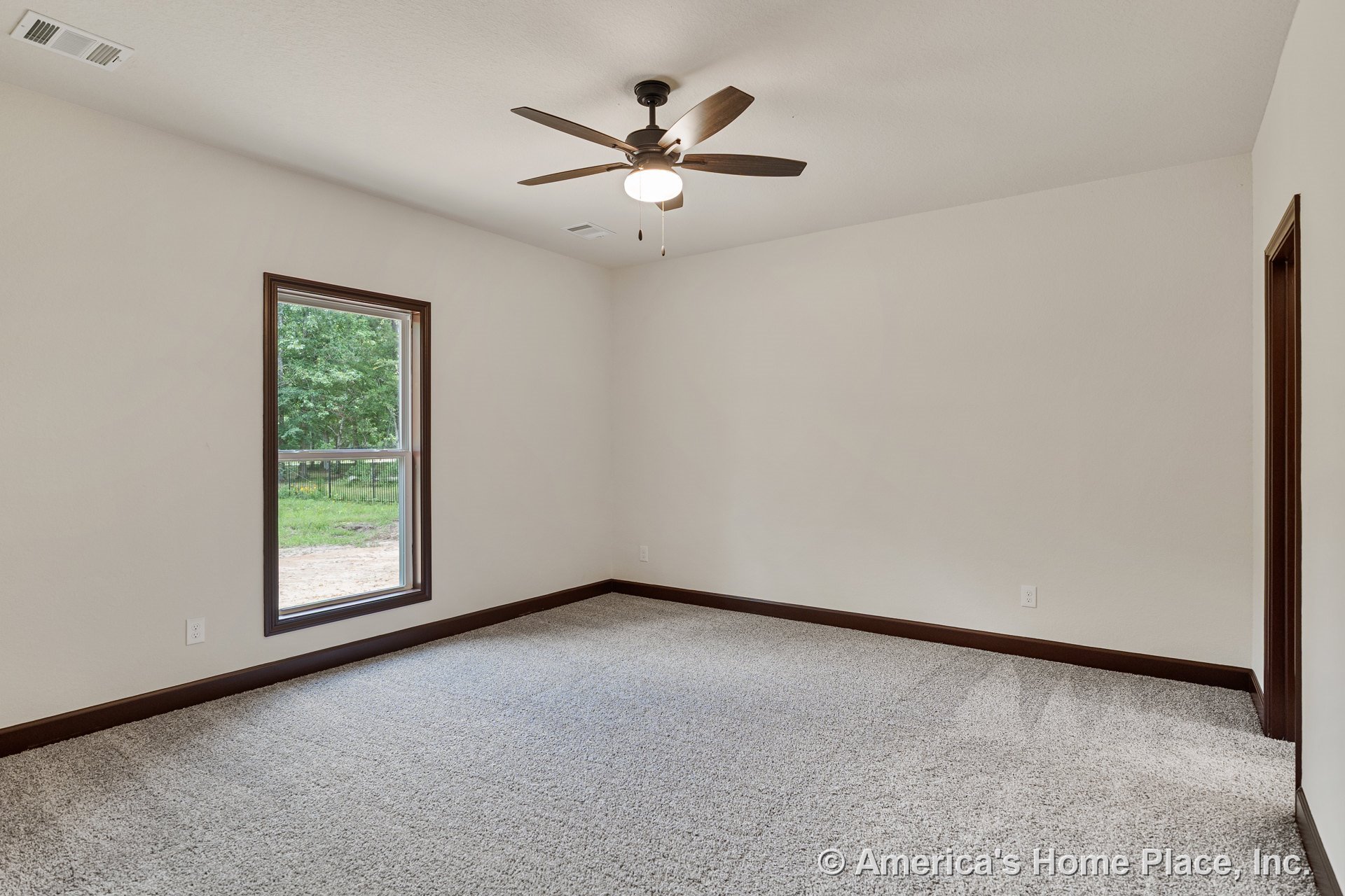 Carpeted bedroom with ceiling fan and light fixture, single window framed in wood trim, neutral walls, wood baseboards, and doorway allowing natural light.