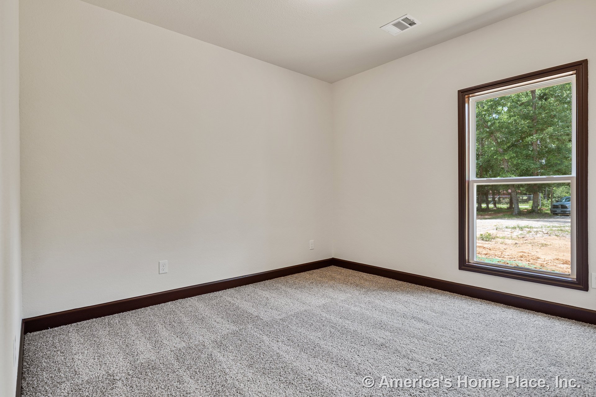 Carpeted bedroom with large window framed by dark wood trim, light neutral walls, matching wood baseboards, ceiling vent, electrical outlet, and rectangular layout.
