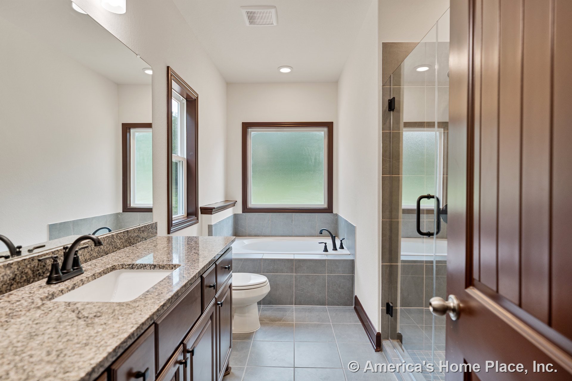 Granite vanity with undermount sink and dark wood cabinets, tiled walk-in shower, soaking tub with tile surround beneath frosted privacy window, recessed ceiling lighting, large
