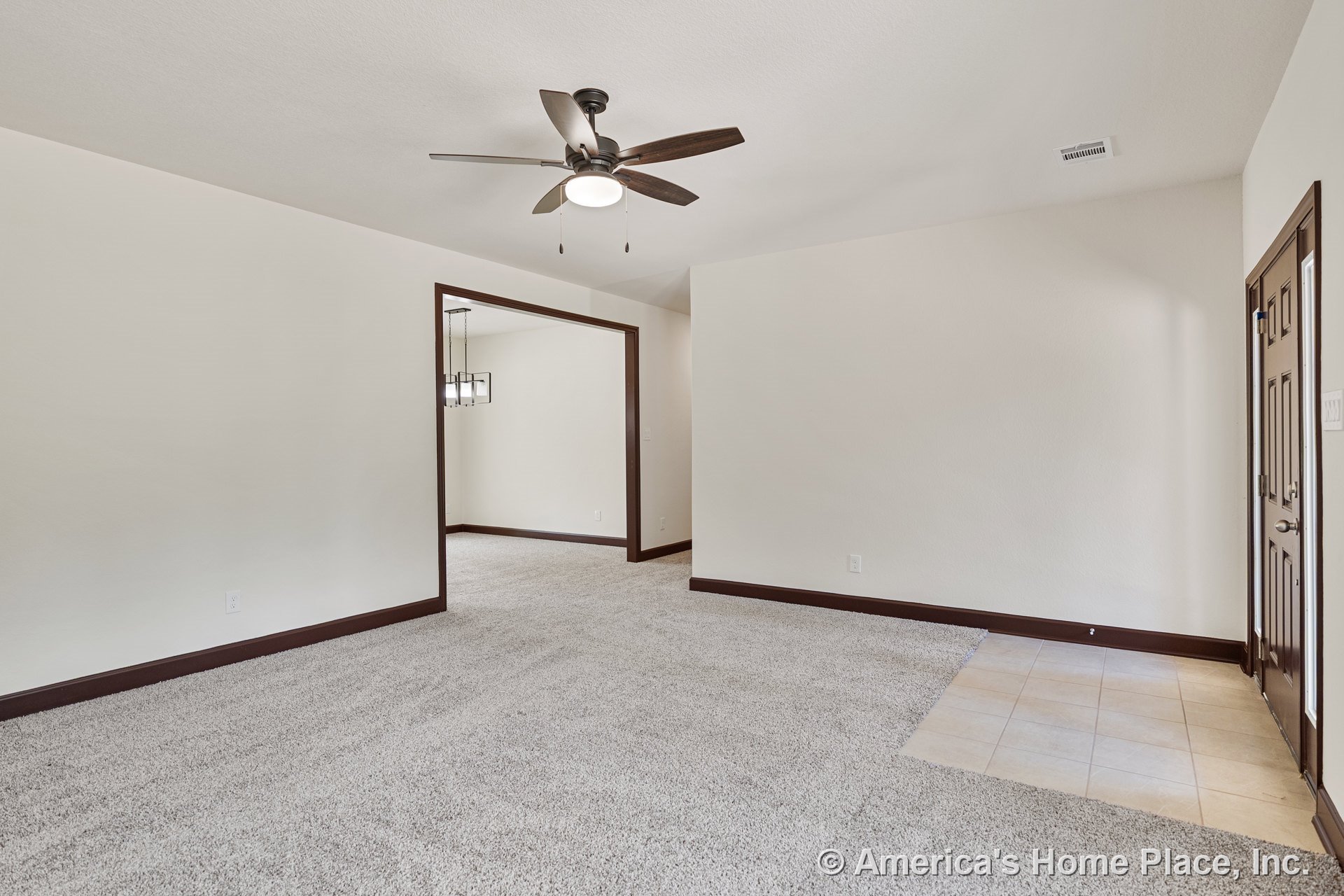 Carpeted living room with ceiling fan and light fixture, dark wood trim around doors and baseboards, tile entryway, open doorway to adjacent room, and neutral wall color.