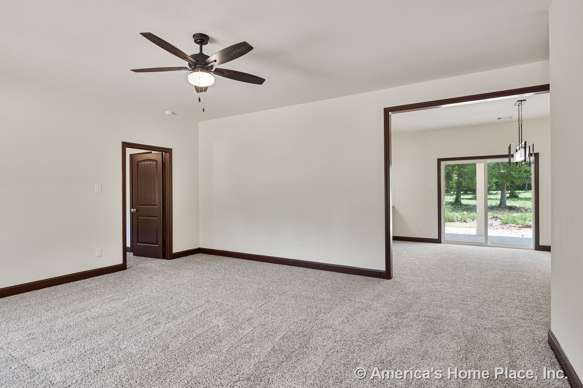 Carpeted living room with dark wood trim, ceiling fan with light, open doorway leading to dining area featuring large sliding glass doors and rectangular chandelier; neutral walls