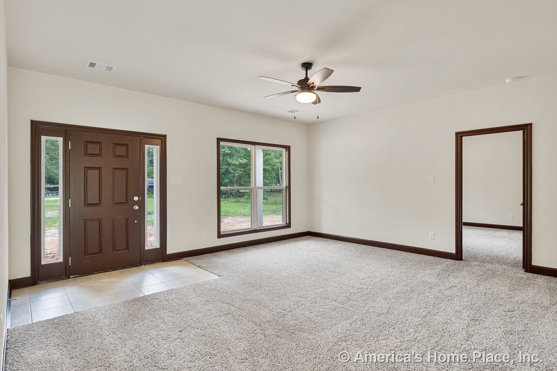 Carpeted living room with large windows framed in dark wood trim, ceiling fan with light fixture, neutral walls, tile entryway, and brown front door with sidelights.