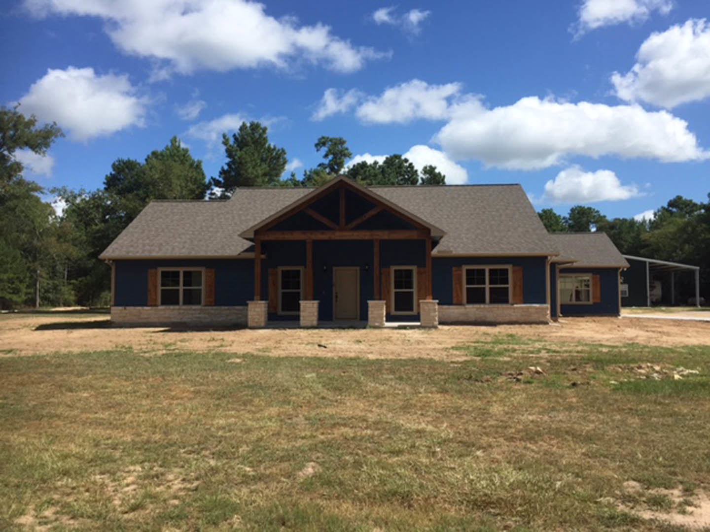 Blue-walled house with white roof and white-framed windows, white front door, expansive grassy yard, mature trees behind the roof, partly cloudy sky overhead