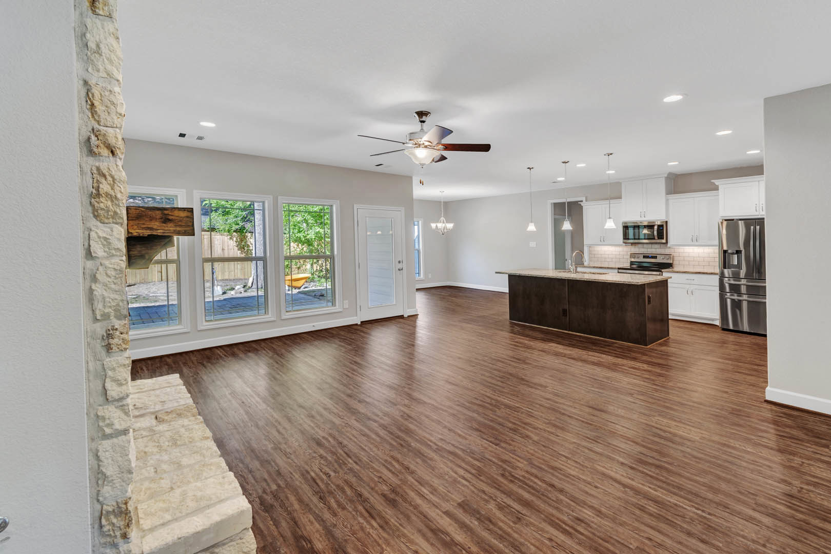 Spacious open floor plan featuring hardwood floors, modern kitchen with stainless steel refrigerator and wood cabinetry, stone fireplace, white glass-paneled door, and large window