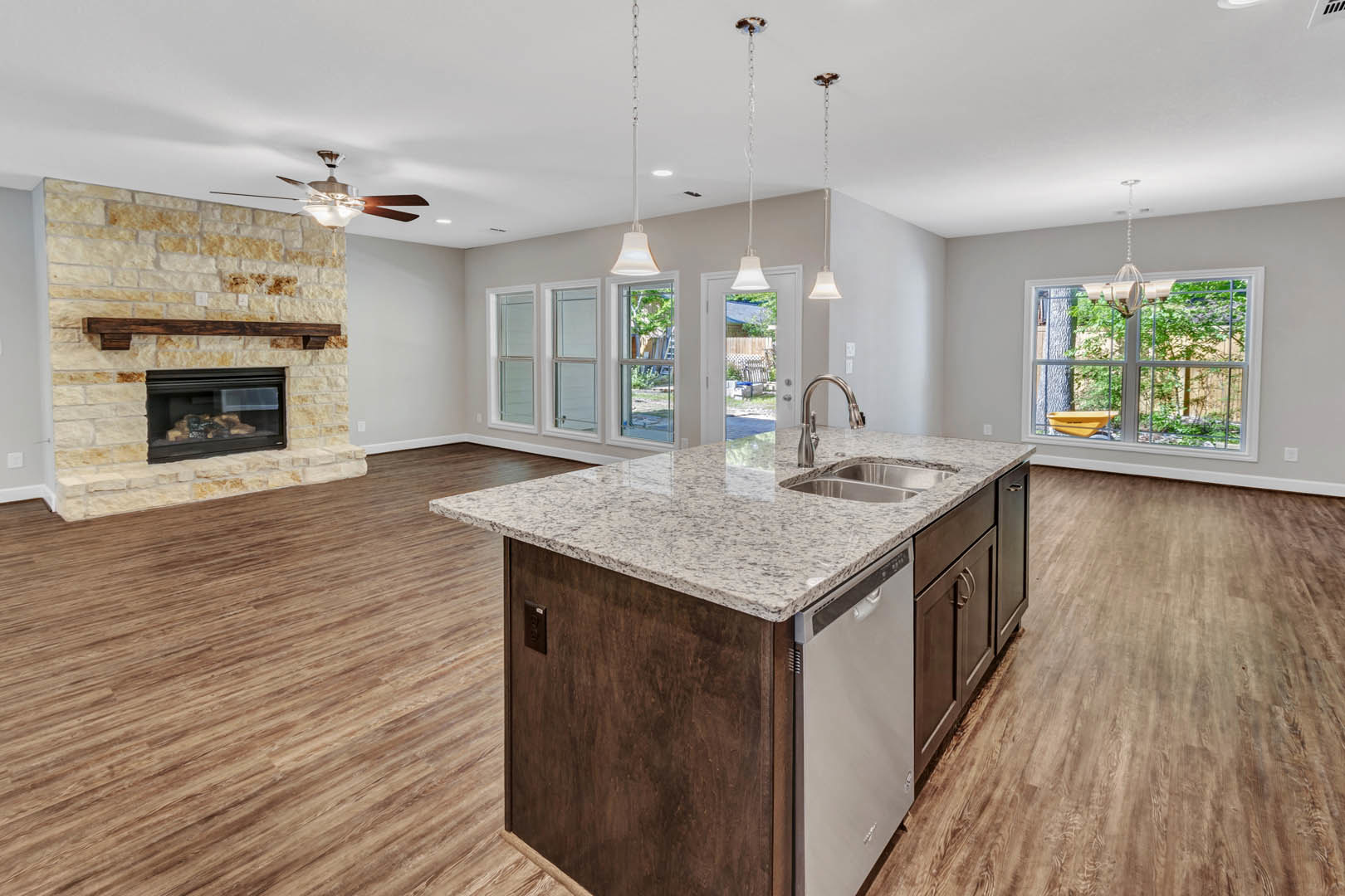 Open-concept kitchen and living room featuring stone fireplace, large window with wheelbarrow visible outside, kitchen island with built-in sink and dishwasher, wood cabinetry