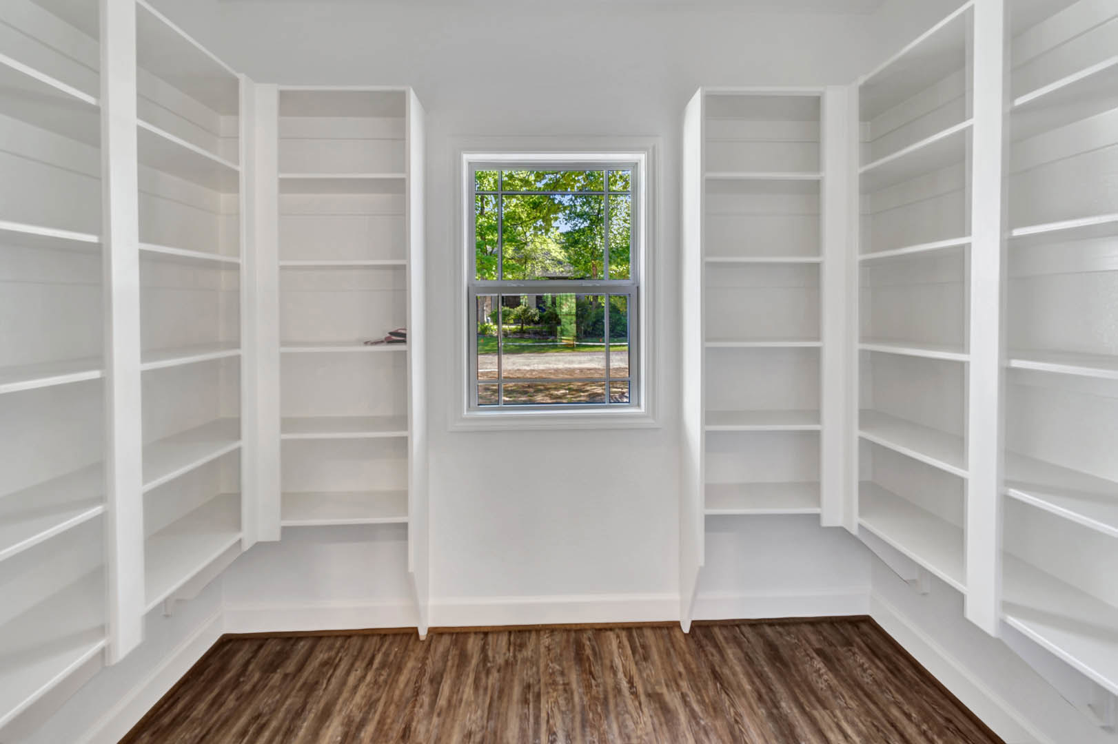 Built-in white shelving along a wood-paneled wall, large window overlooking yard and trees, hardwood flooring