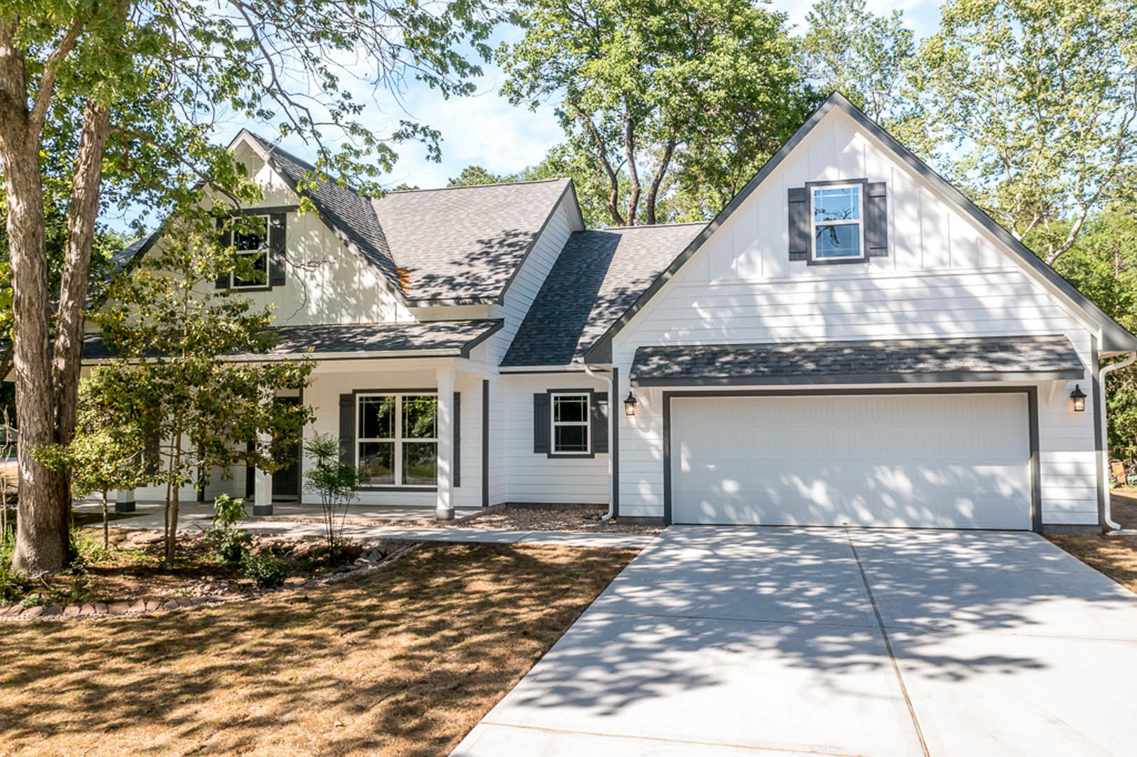 Modern house with white garage door, black roof, concrete driveway shaded by mature trees, white-framed window, landscaped front yard, and blue sky overhead