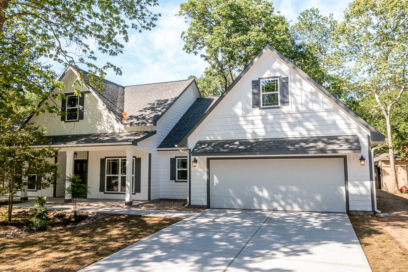Two-story home with white siding, concrete driveway leading to a white garage door, front porch, large windows with white frames, mature trees and landscaping in the yard