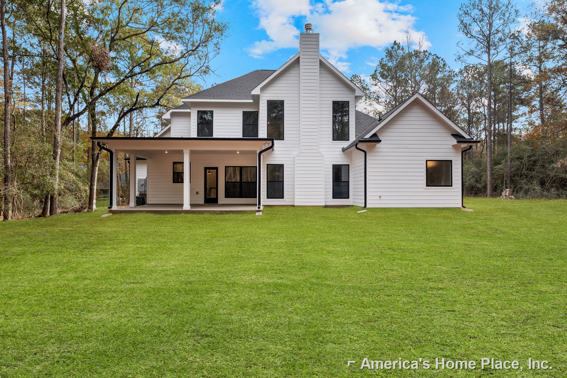 White farmhouse-style home with black-framed windows, white siding, prominent chimney, and expansive green lawn under a partly cloudy sky
