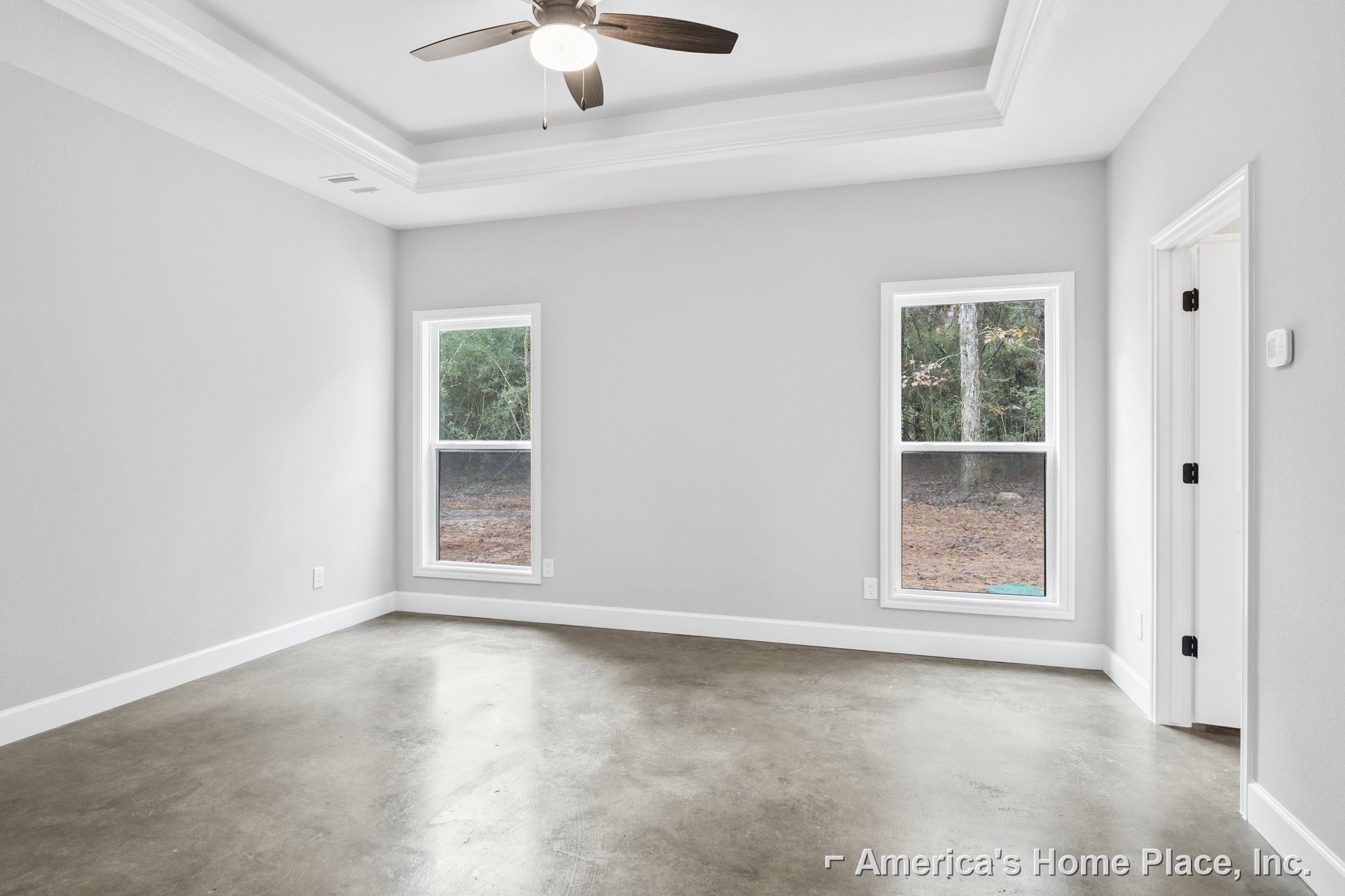 Ceiling fan with light fixture above concrete floor, white baseboard trim, large windows showing leafy trees outside