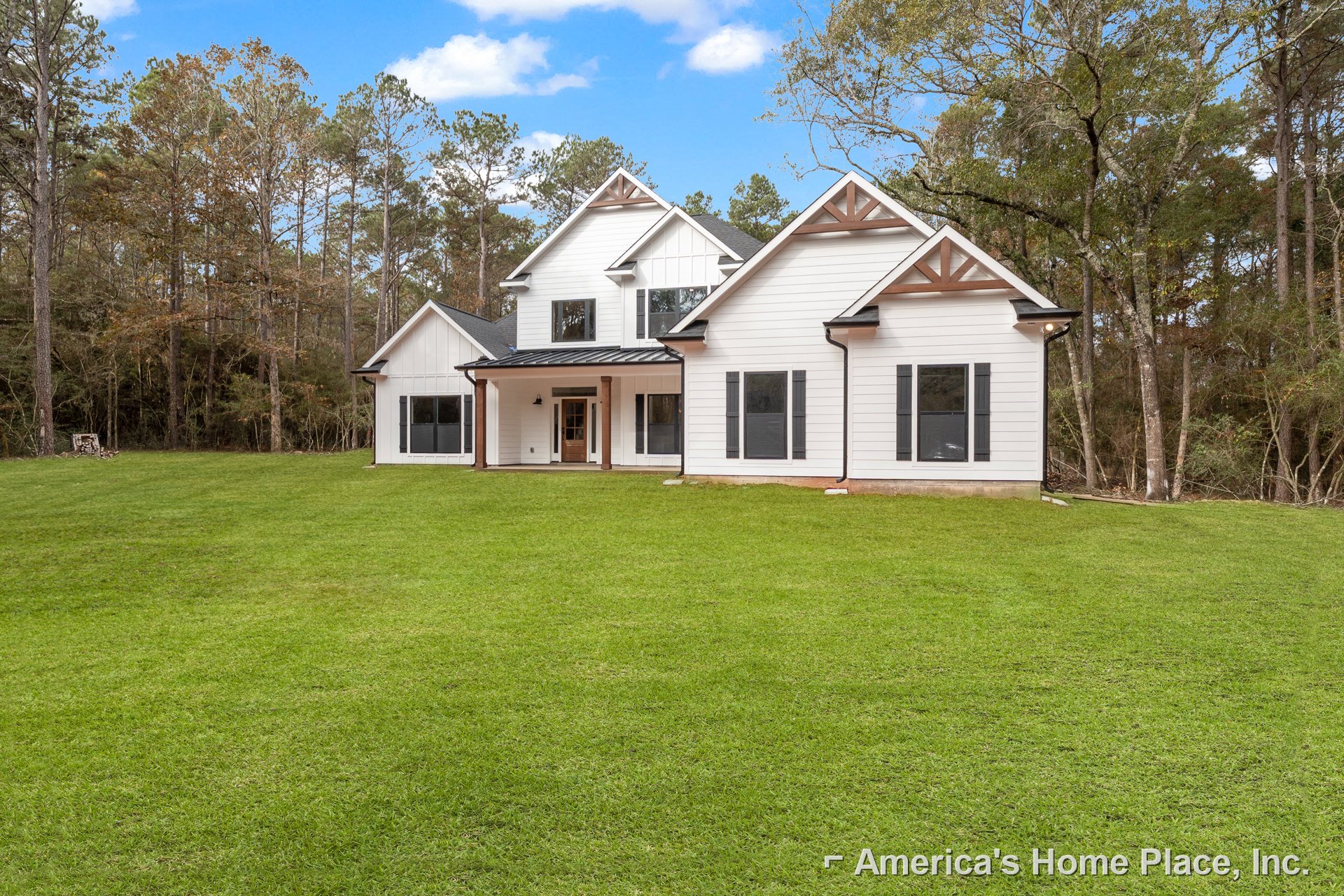 White farmhouse with black roof, front lawn of green grass, large windows reflecting trees and parked car, entry door with glass panel, partly cloudy sky overhead