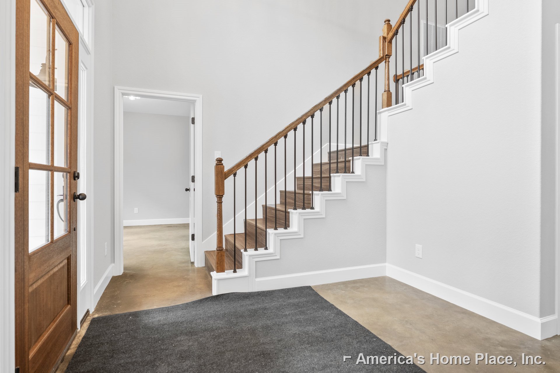 Wood and metal staircase with black handrail, white door with black handle, patterned rug on light hardwood floor, white walls
