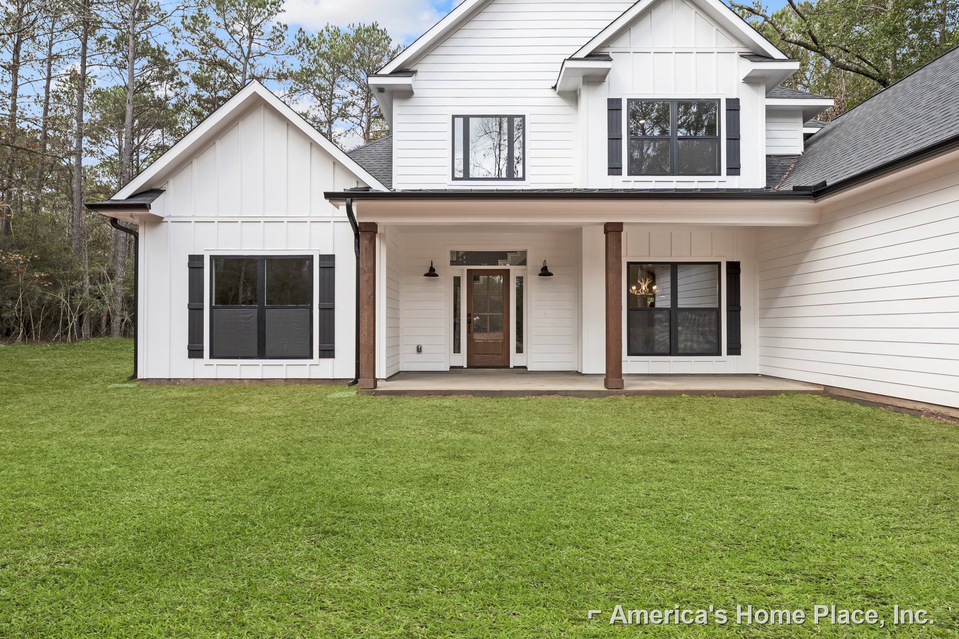 White siding house with black-framed windows, front porch with glass door reflecting chandelier, green lawn, and trees reflected in window.