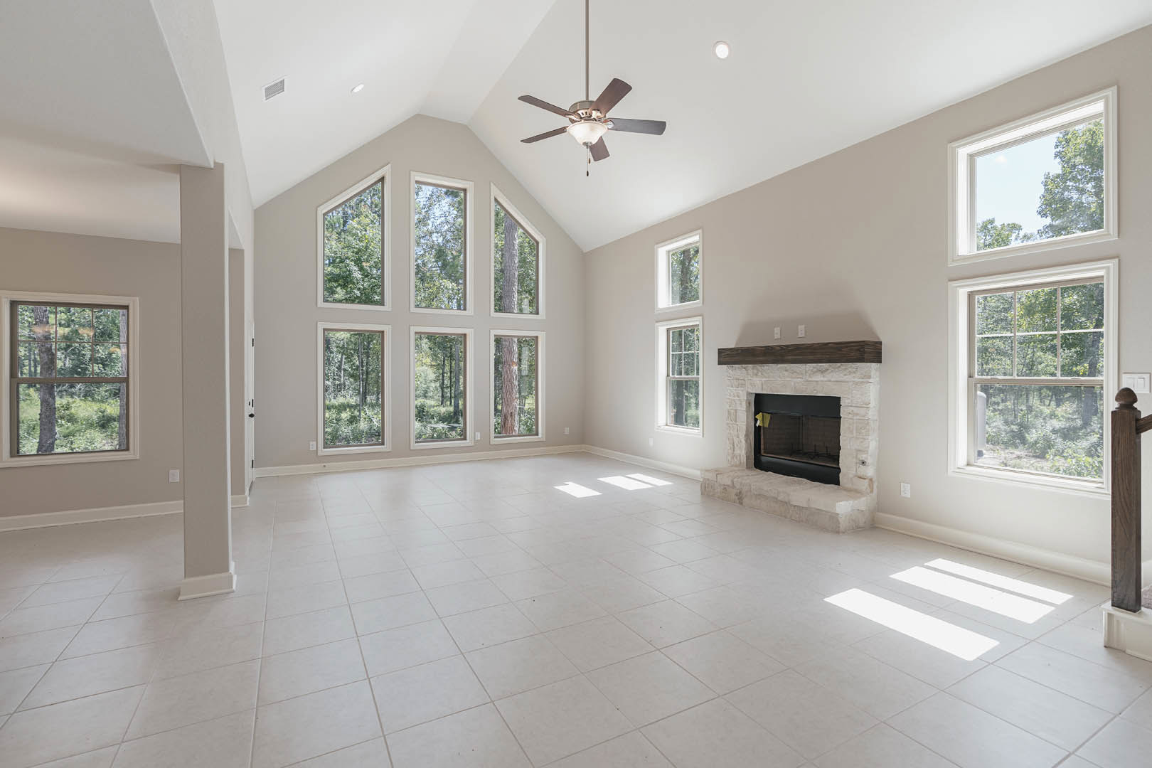 Spacious living room with white tile flooring, black fireplace featuring a wood mantle and yellow post-it note, ceiling fan with light fixture, large windows overlooking forest