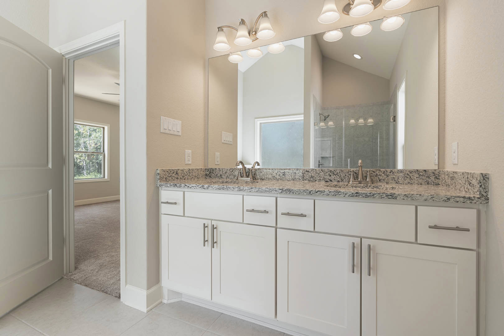 Bathroom with expansive vanity topped with light stone countertop, wide mirror above, white cabinetry, chrome faucet, and window framing leafy trees outside
