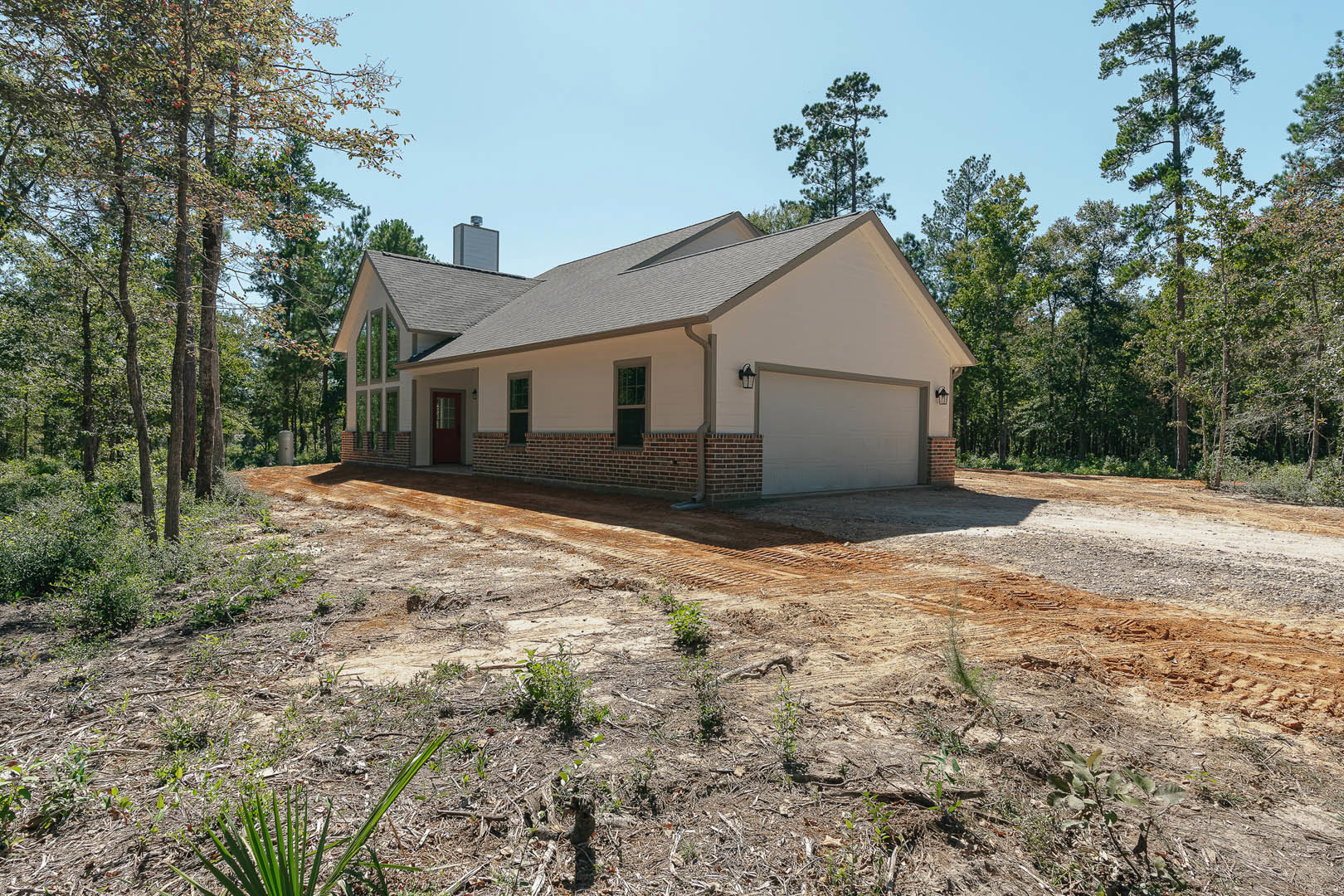 White house with attached garage, dirt driveway bordered by green plants, mature trees in background, brick structure partially visible