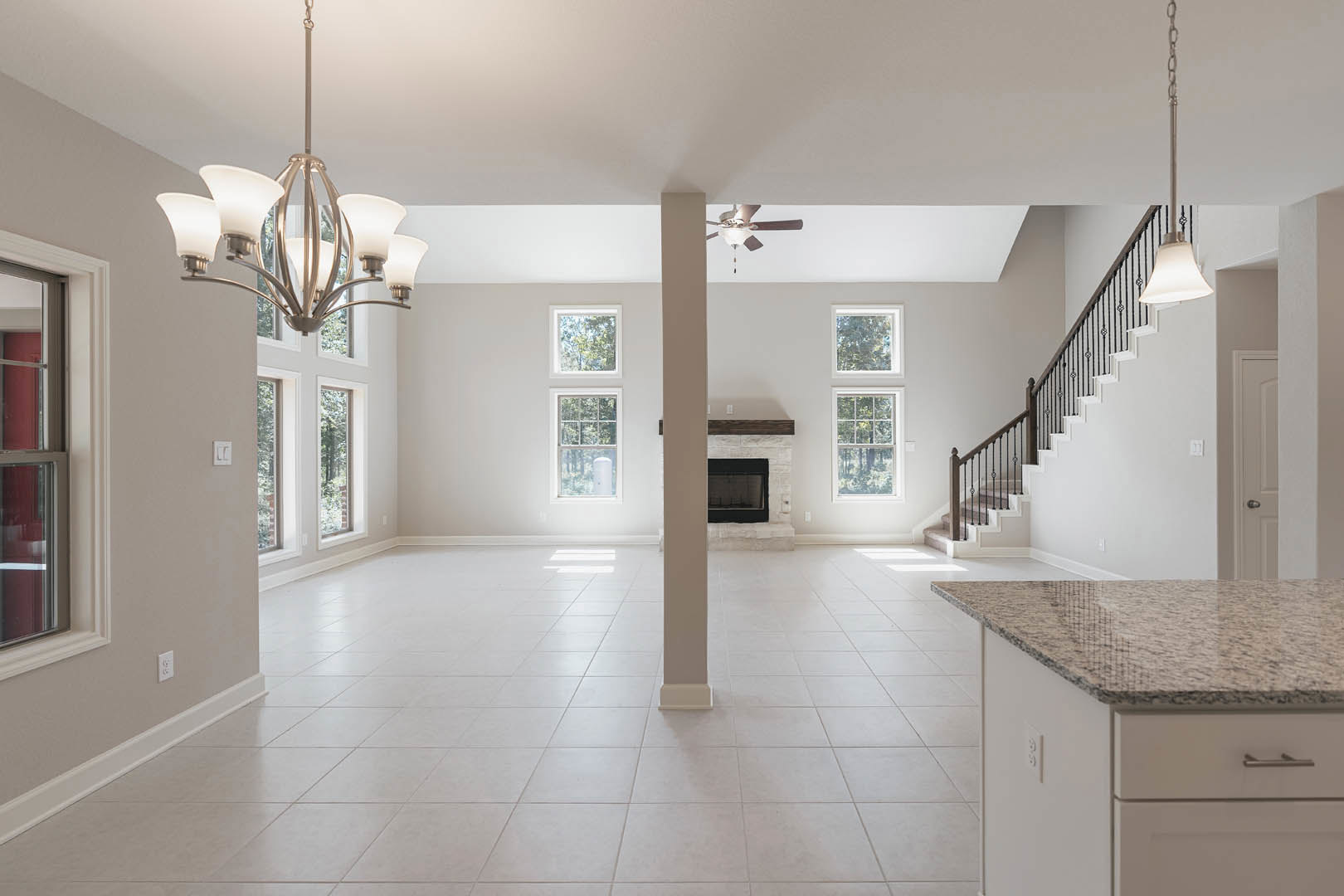 Spacious white living area featuring a modern staircase, sleek fireplace, white cabinetry with stone countertop, decorative pillar, ceiling fan, and black-framed glass window.