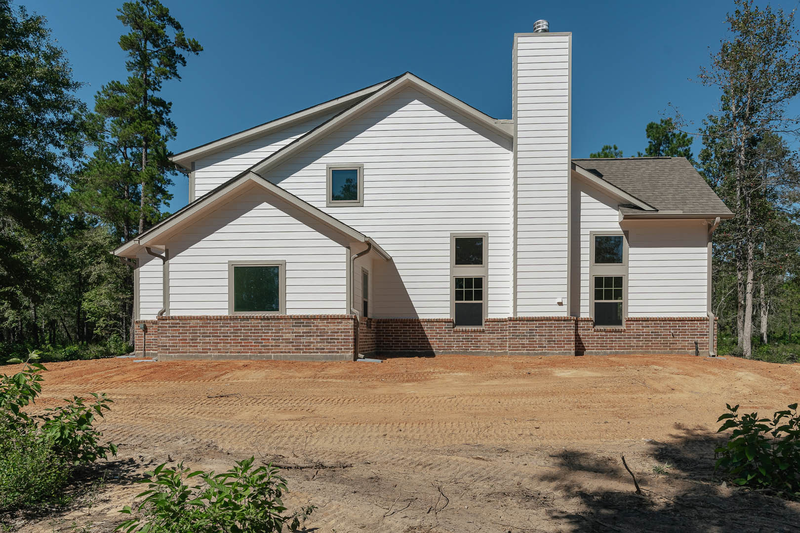 White cottage with brick walls and white-framed windows, dirt road with tire tracks in foreground, tree and American Gothic House visible in background under open sky.