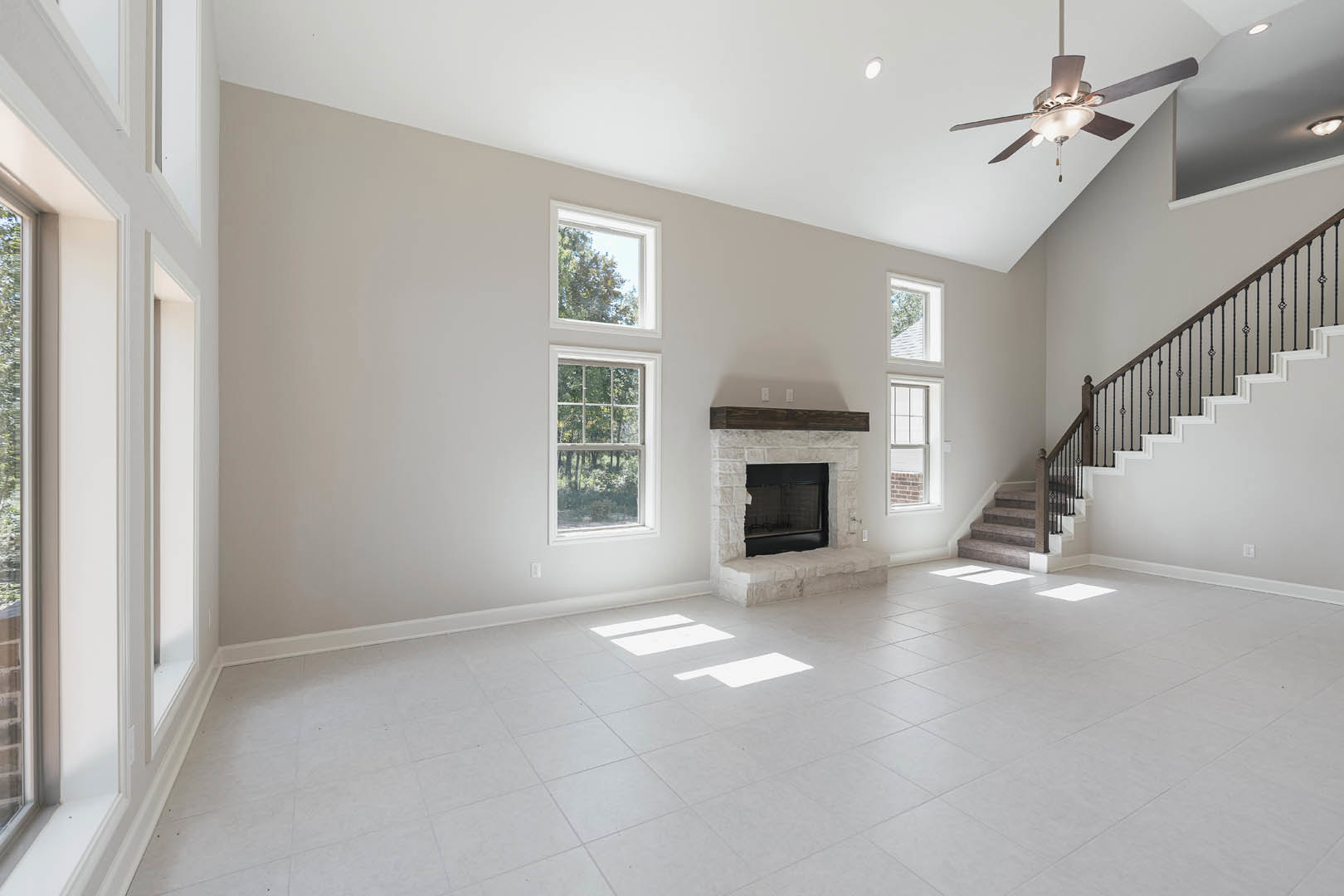 Spacious living area featuring white tile flooring, a central fireplace with wood mantle, staircase with wooden railing, ceiling fan with light fixture, and large window