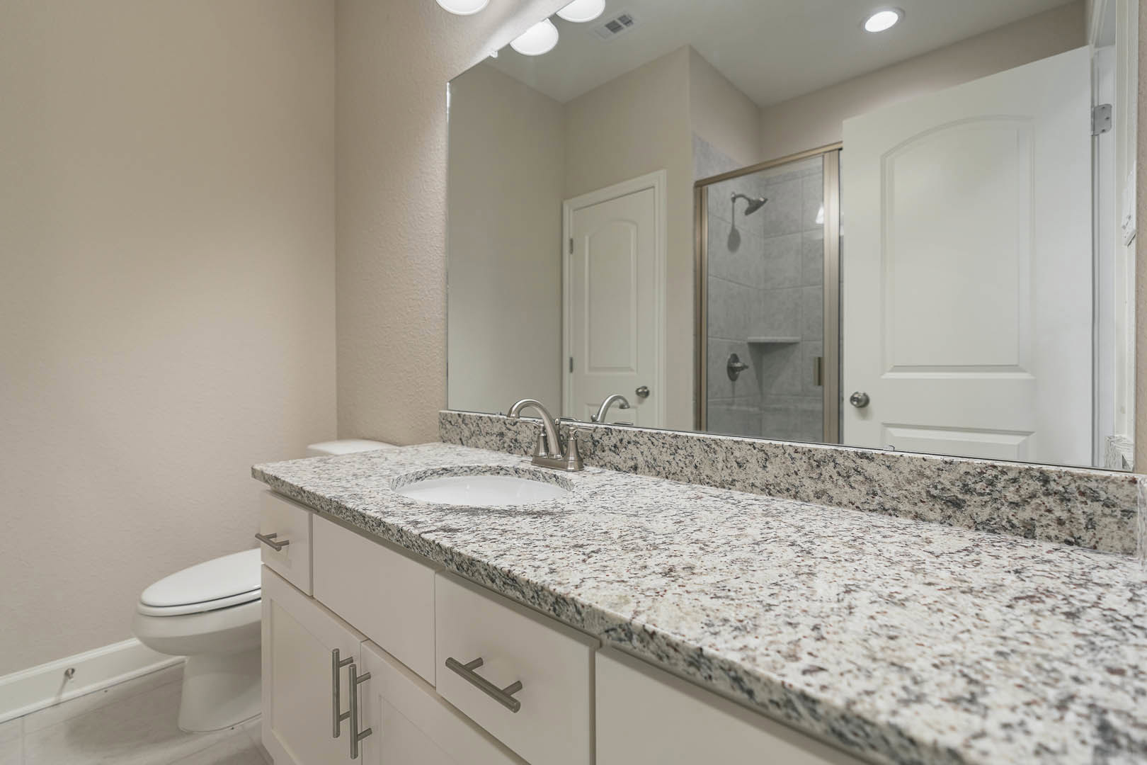 Marble bathroom countertop with undermount sink and chrome faucet, white cabinetry, tiled shower with glass door, and toilet visible in the background
