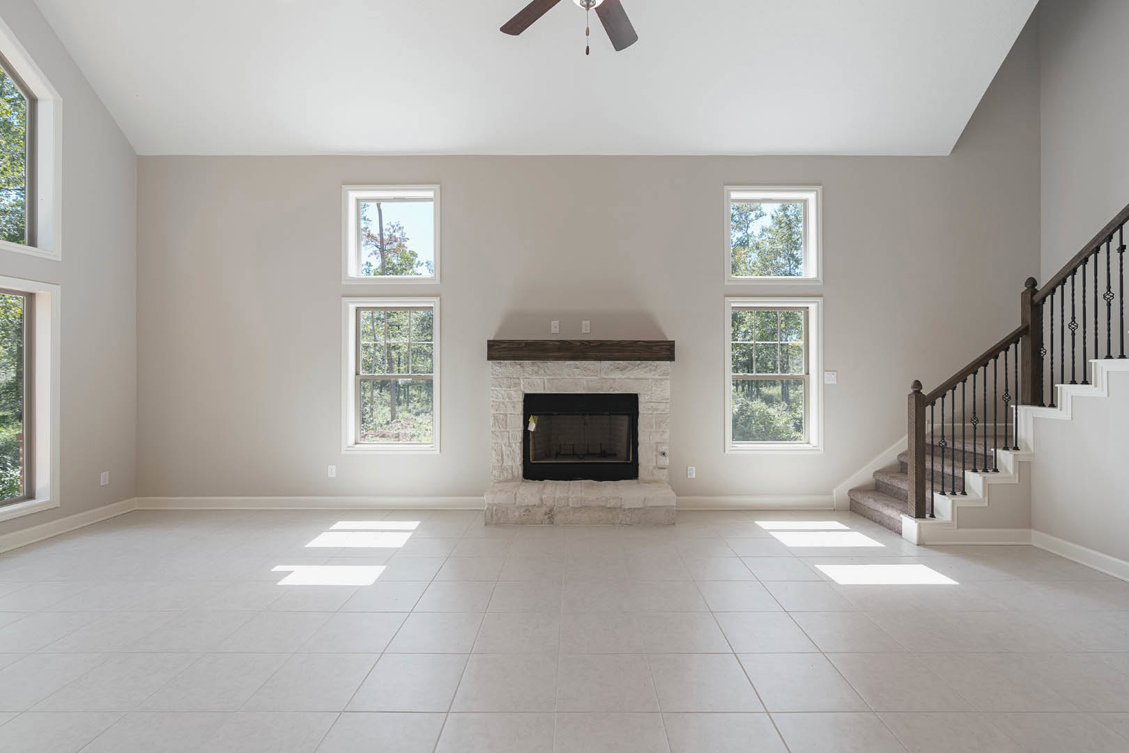 Living room with white tile floor, stone step, wood beam fireplace, ceiling fan with light, staircase, and window showing trees outside