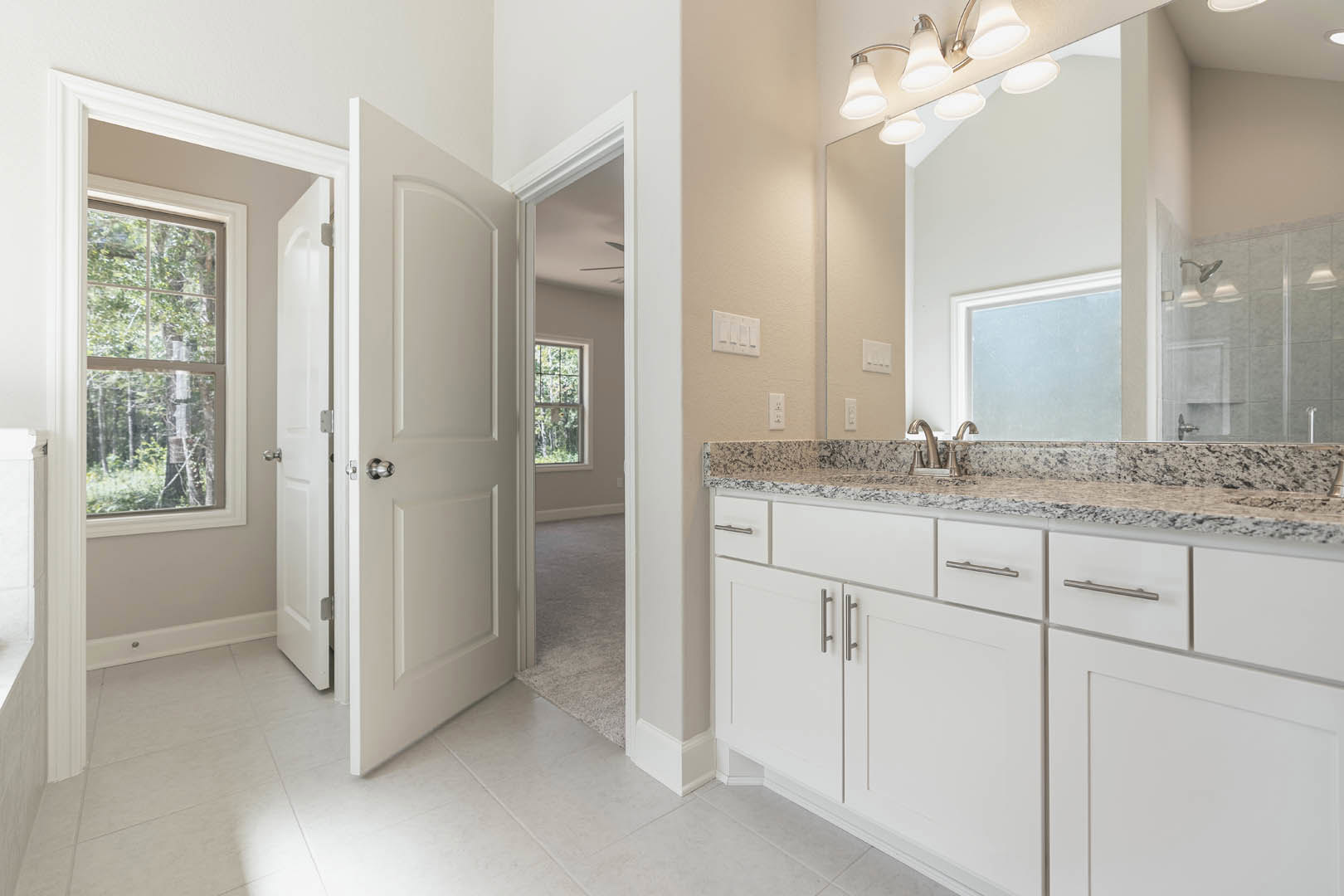 Bathroom with marble countertop, large mirror above sink, white cabinetry, frosted glass window, shower head and faucet, view of trees through window.
