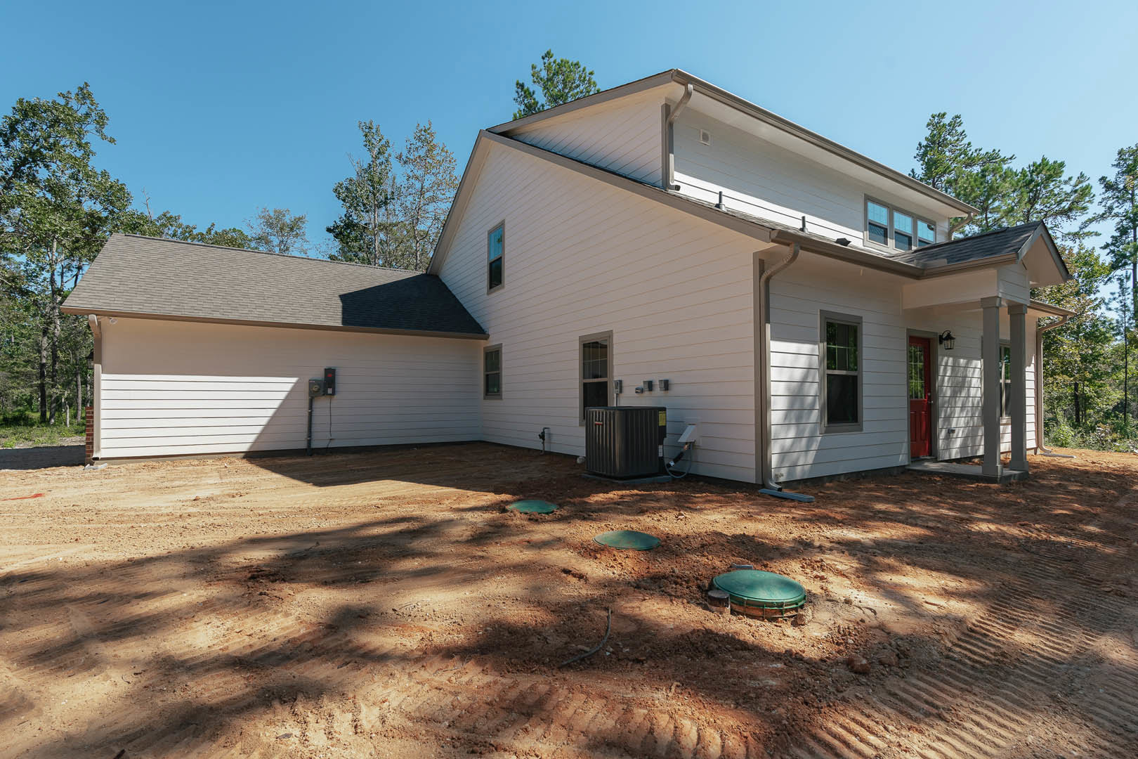 White house with grey roof, black-framed windows, and black front door; dirt yard with patches of green grass, green manhole cover, and trees in the background