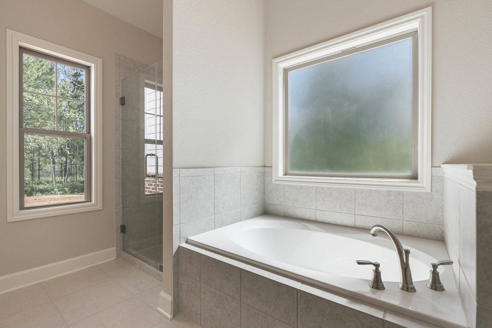 White tile bathroom featuring a freestanding bathtub, glass-enclosed shower with chrome fixtures, frosted window overlooking trees, and modern sink.