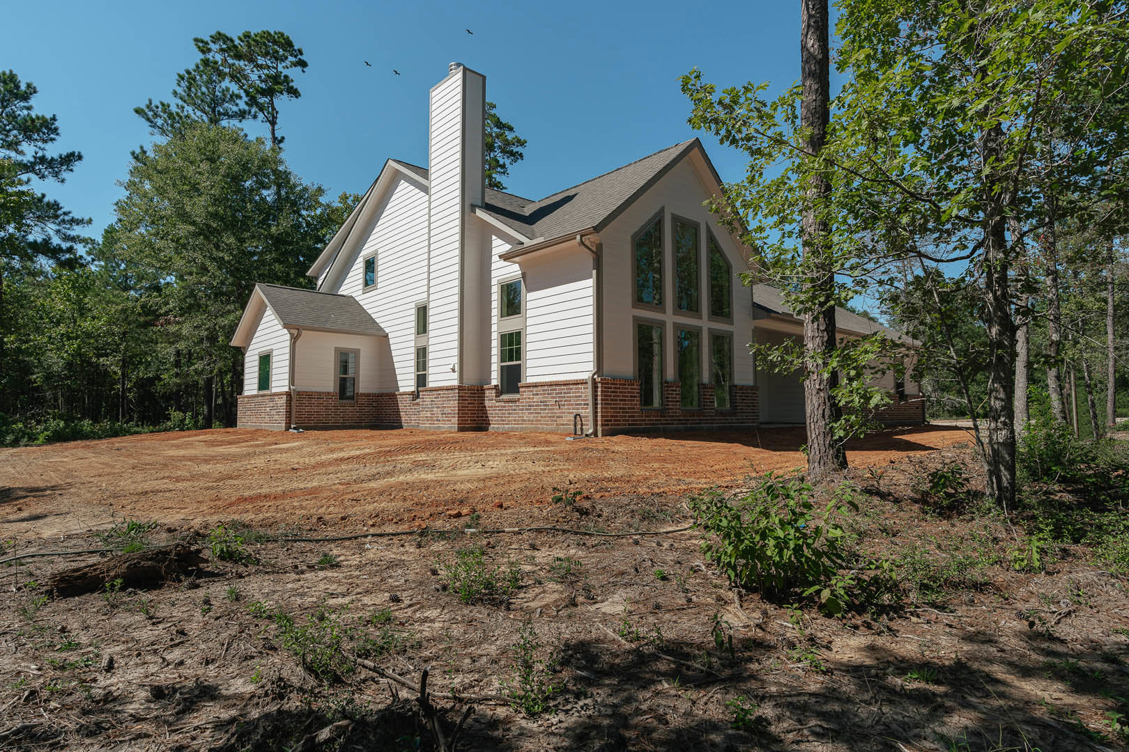 Brick chimney rises above a gabled roof, surrounded by mature trees; windows and siding visible, with a dirt yard and scattered plants in foreground.