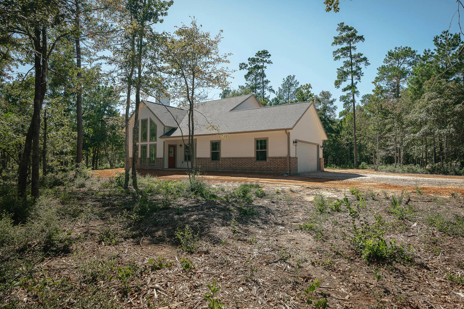 Two-story brick and white stucco house surrounded by mature trees, brown-framed windows, small plants on a dirt patch in front yard