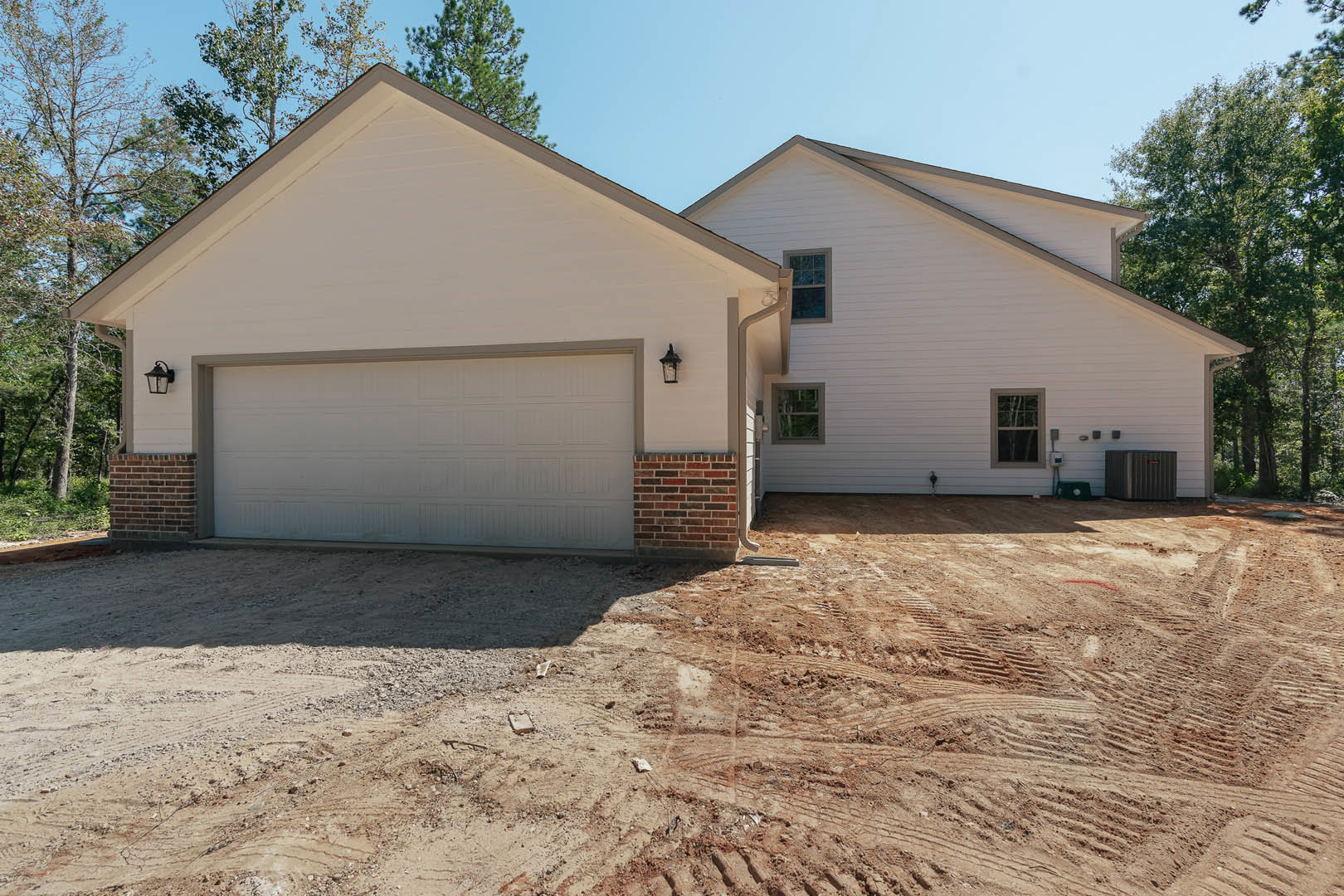 Two-story house with white-framed windows, attached garage with paneled door, dirt driveway showing tire tracks, tile accent wall near window, surrounded by trees under blue sky