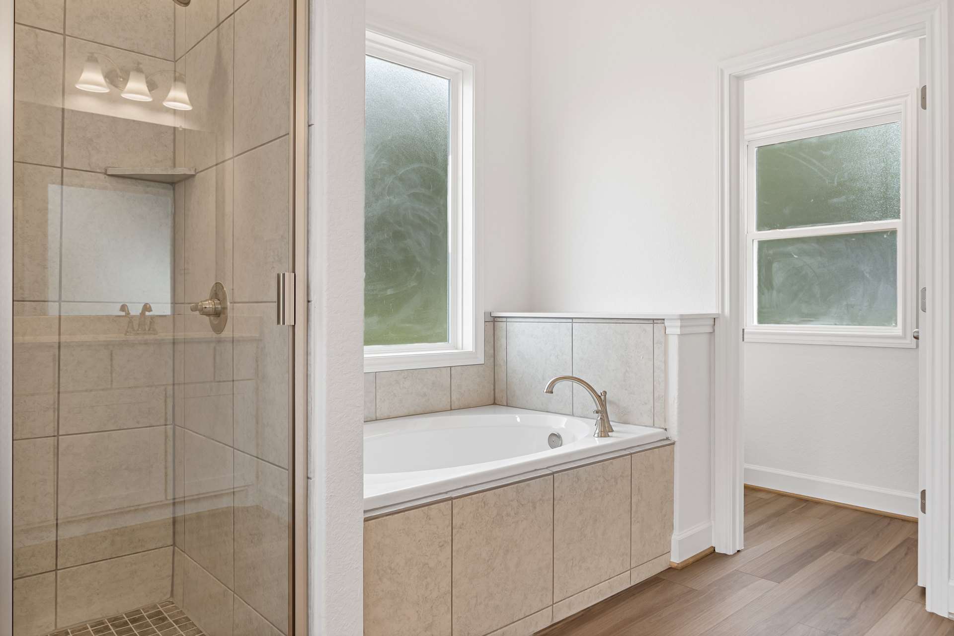 White bathtub with silver faucet beside tiled shower, large window, and modern light fixture in contemporary bathroom.