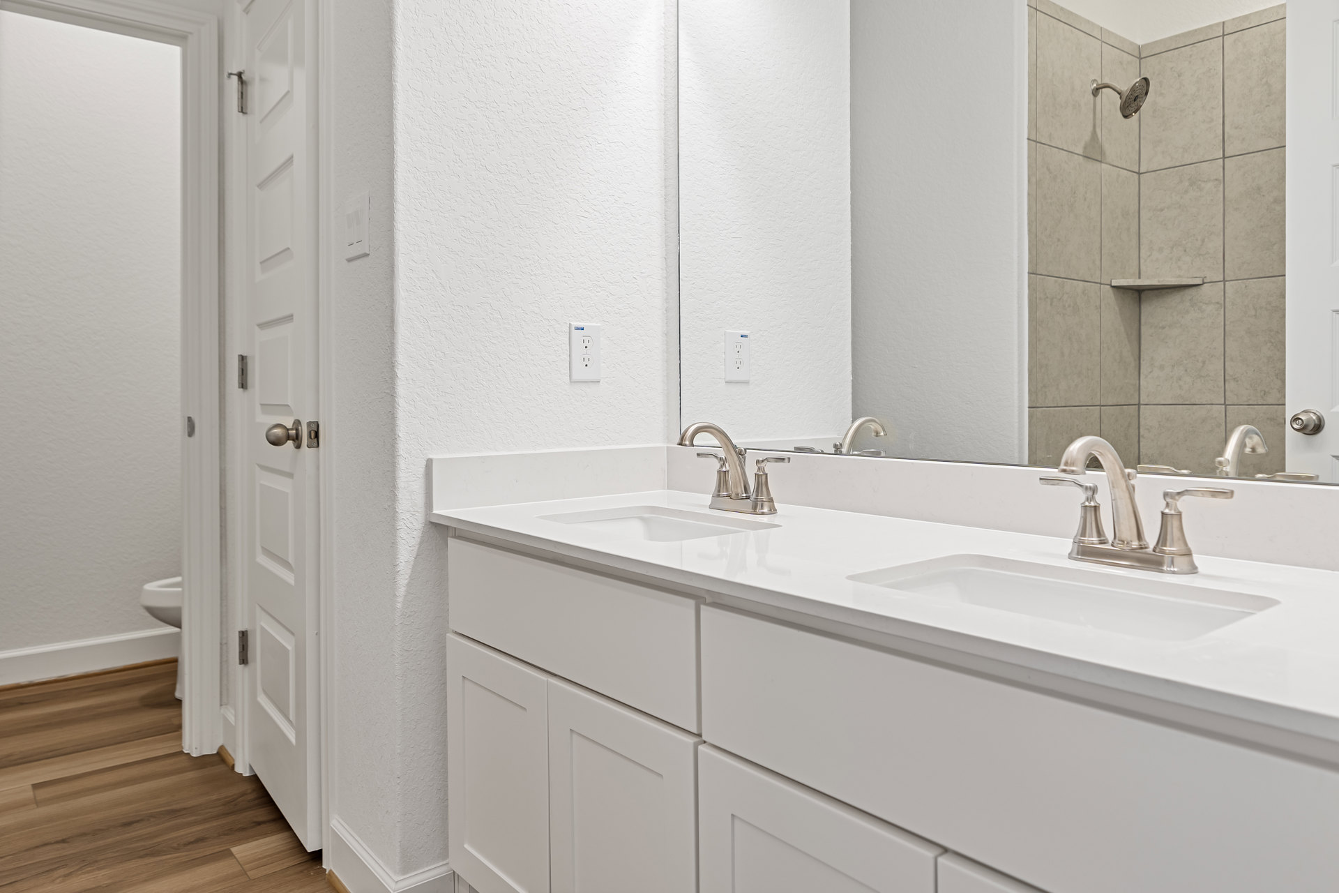 Bathroom with white cabinets and walls, wood flooring with white trim, white countertop and sink, chrome faucet, large mirror above vanity, minimal black accent line on wall