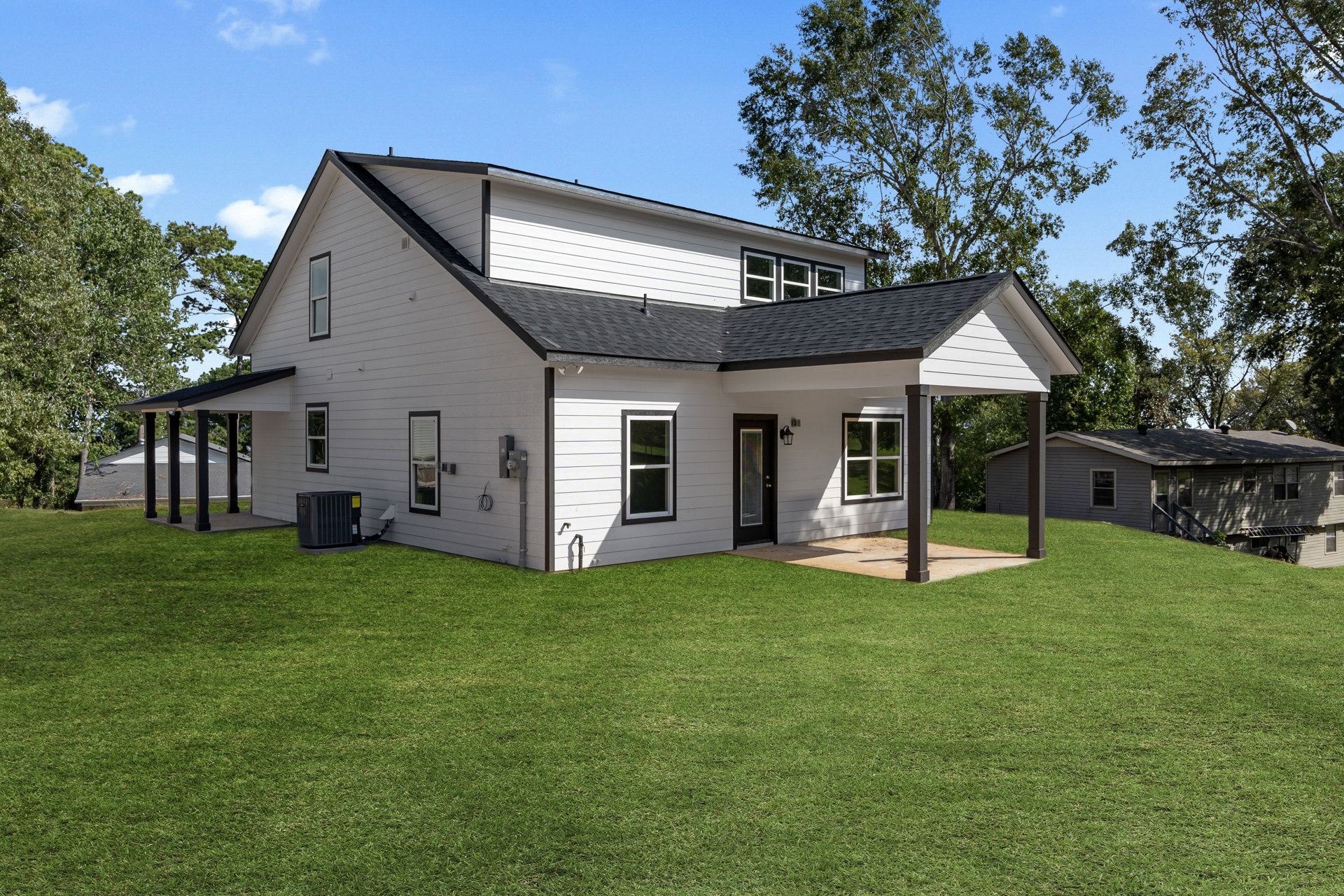 Two-story home with white siding, large windows, and dark roof, surrounded by green lawn and mature trees under a blue sky