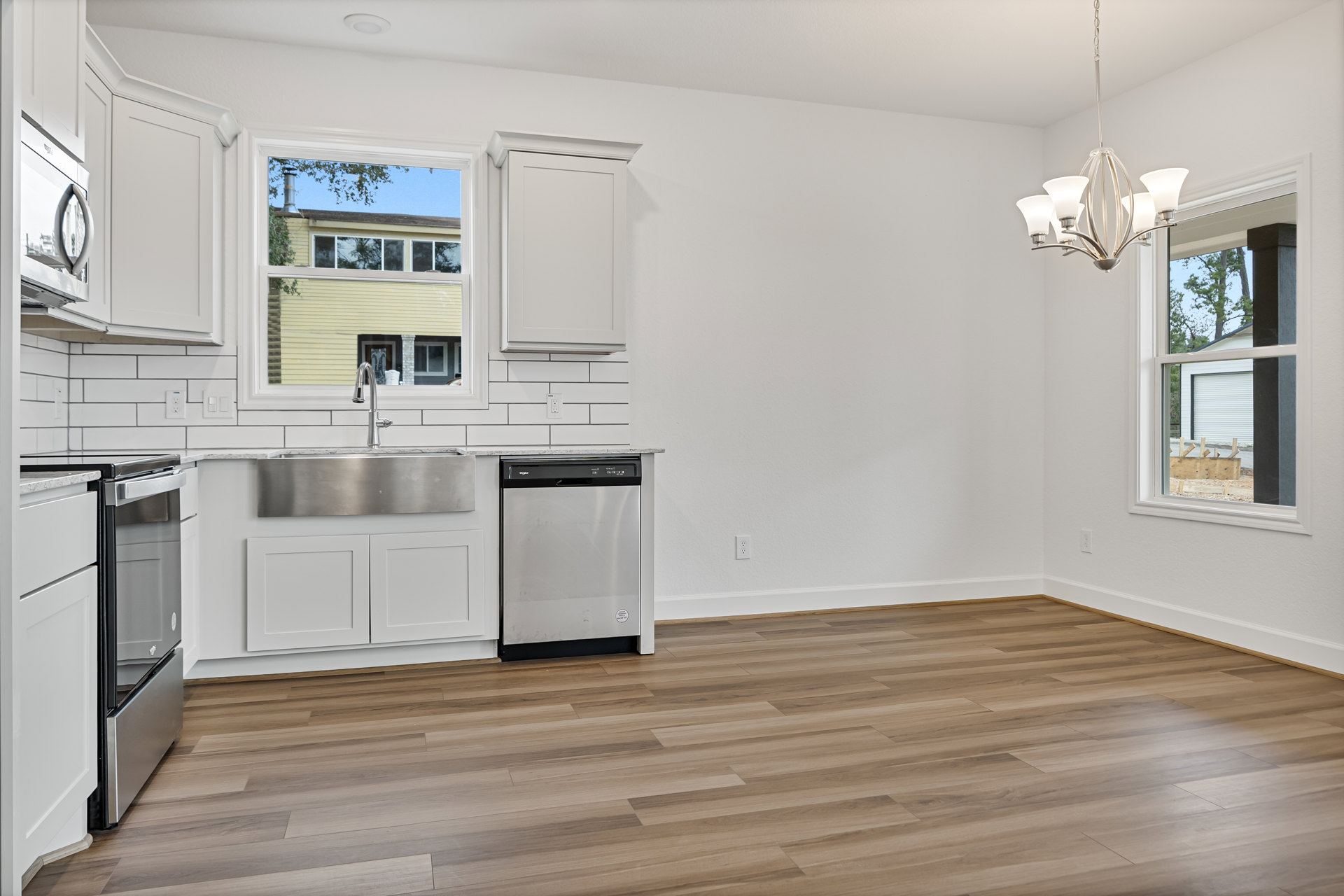 Modern kitchen with wood flooring, white walls, stainless steel refrigerator and dishwasher, white cabinetry, tile backsplash, sink beneath a window, white framed door, and a