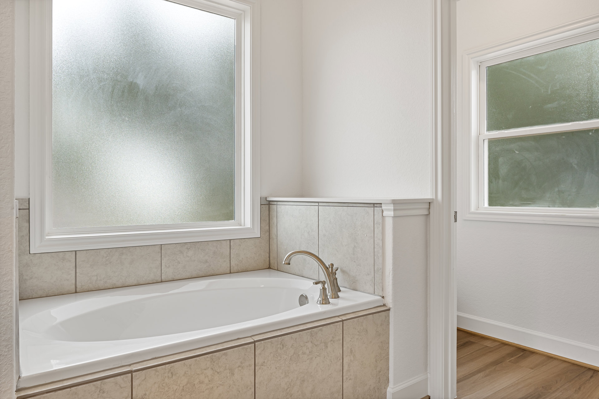 Freestanding white bathtub with chrome faucet beneath frosted window, surrounded by light gray tile walls in modern bathroom