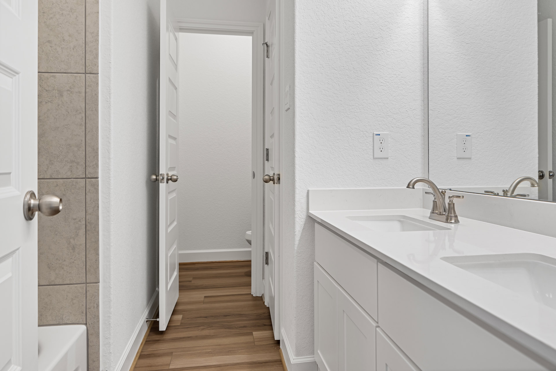 Bathroom with white countertop featuring two sinks, wood flooring, white door with silver handle, and close-up of outlet and door knob