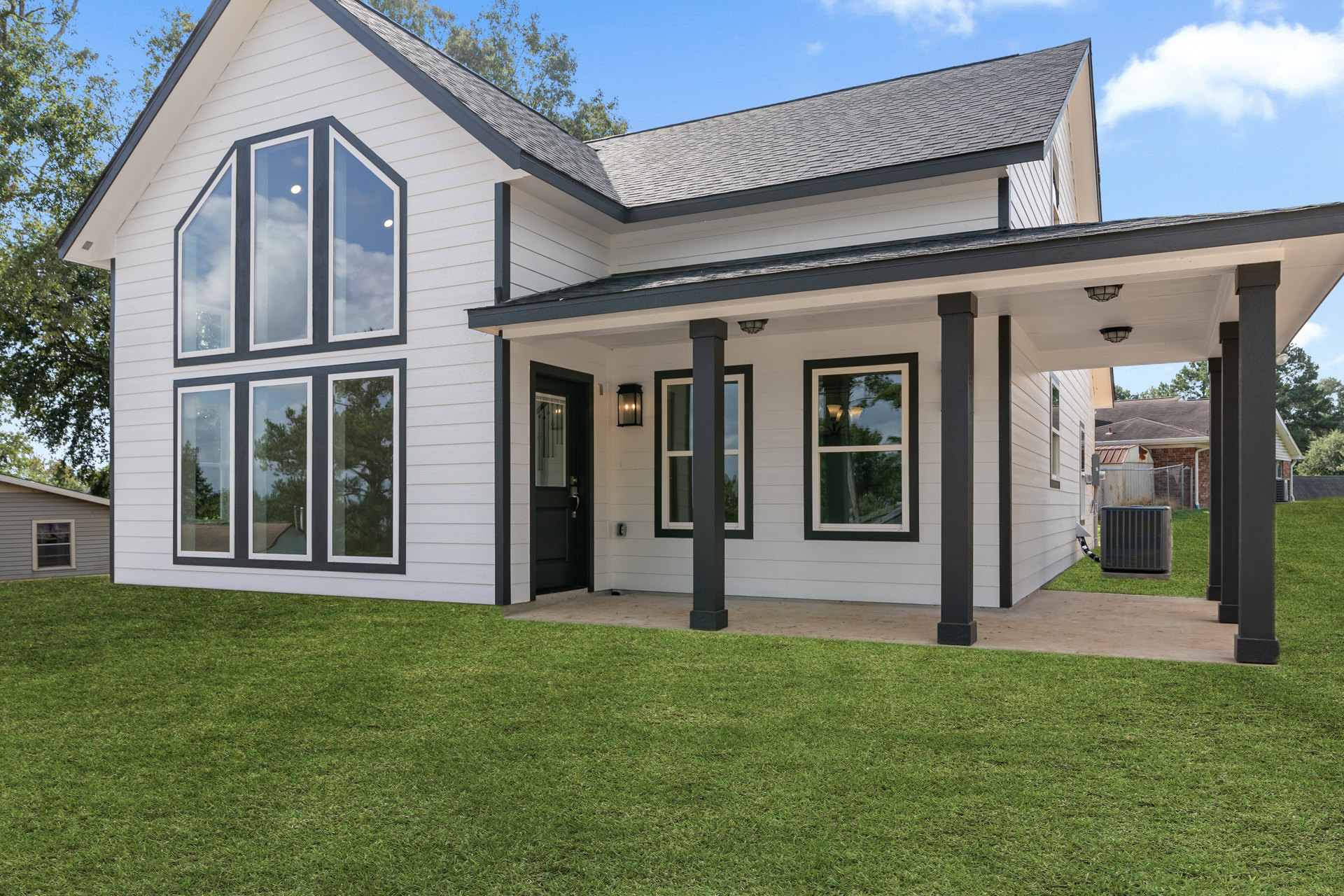 White siding house with multiple windows, manicured green lawn, porch area, black post, and large grey utility box with attached hose; tree reflections visible in window glass.