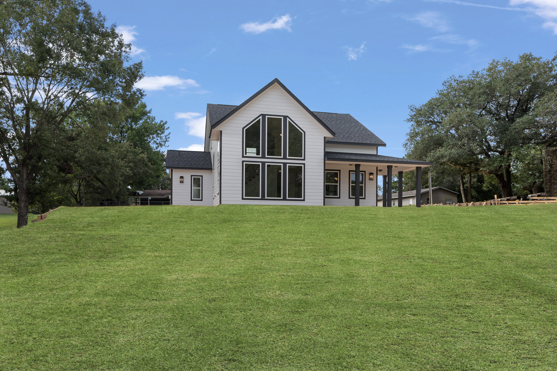 Modern farmhouse with expansive green lawn, large front windows, mature leafy tree beside the house, cloudy sky overhead