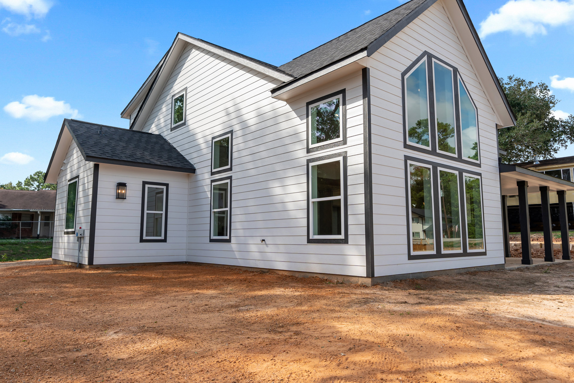 White siding house with black trim, white-framed windows reflecting trees, dirt driveway and ground, gray shingle roof under cloudy sky