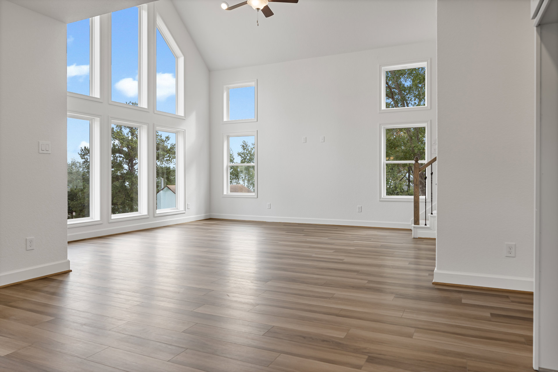 Spacious room with wide plank hardwood floors, multiple large windows showing blue sky and leafy trees, white walls, and natural light filling the space