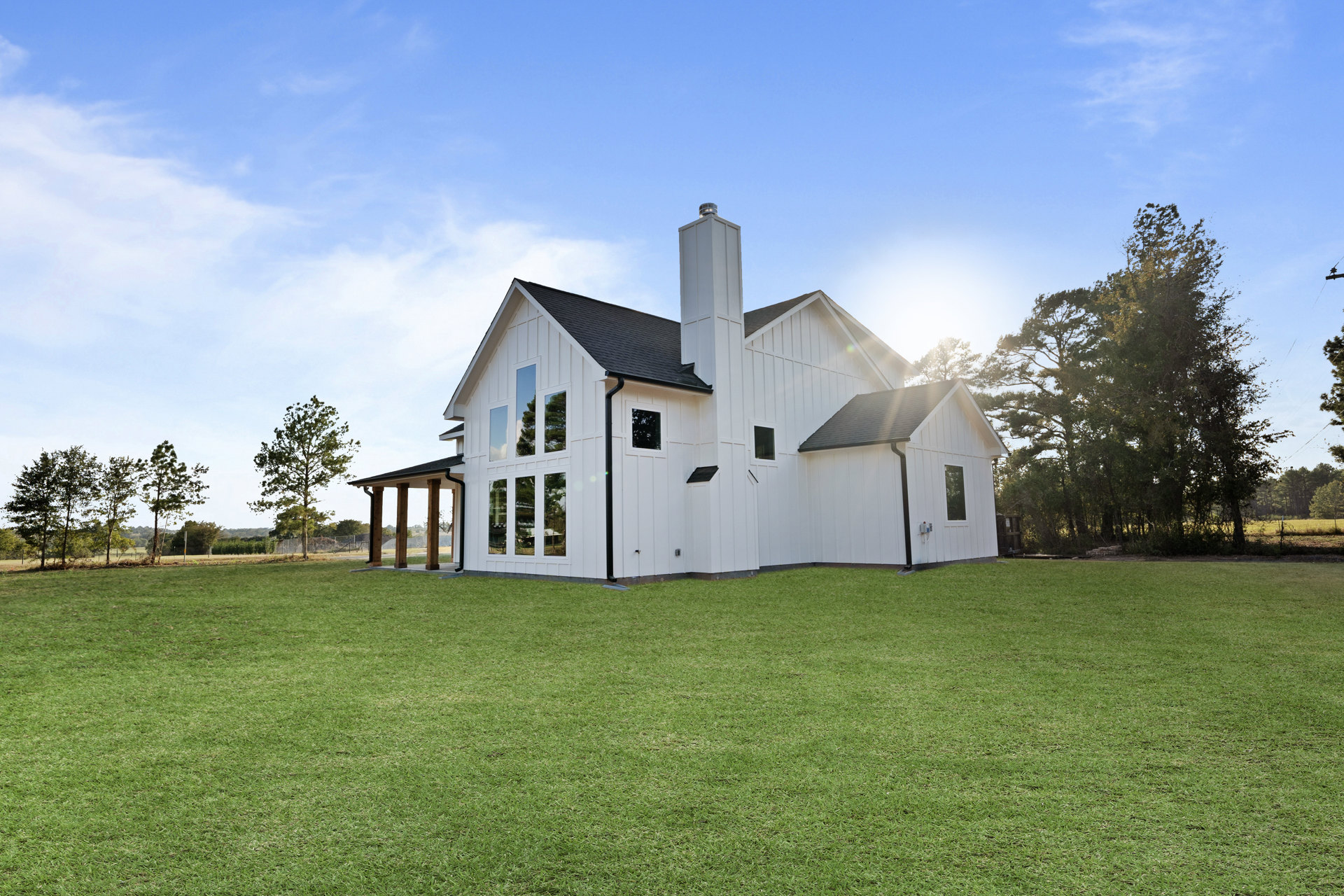 White farmhouse with covered porch, white pillars, brick chimney, expansive green lawn, and tall leafy tree under partly cloudy sky