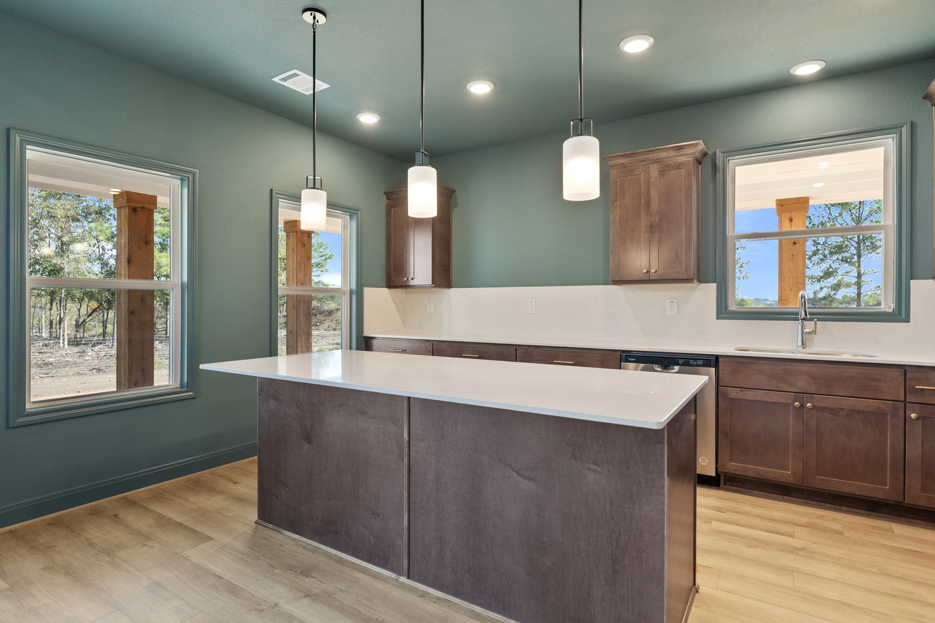 Modern kitchen featuring a large white island with built-in sink, pendant lights overhead, white countertops, wood cabinetry, tile flooring, and windows offering views of trees and