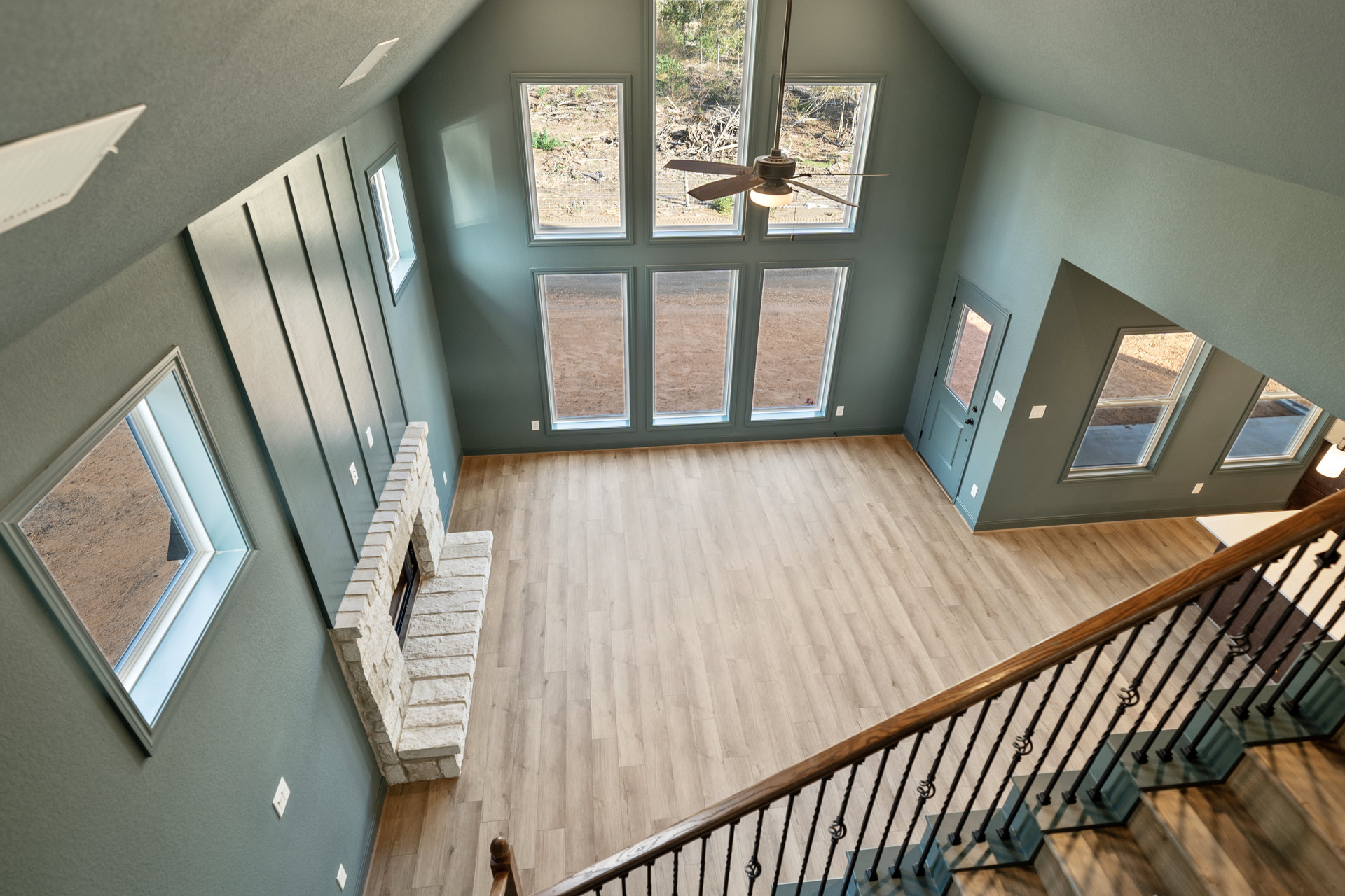 Wood-floored living room with central fireplace, ceiling fan, staircase with wood handrail, and multiple windows overlooking a fenced yard with dirt and plants.