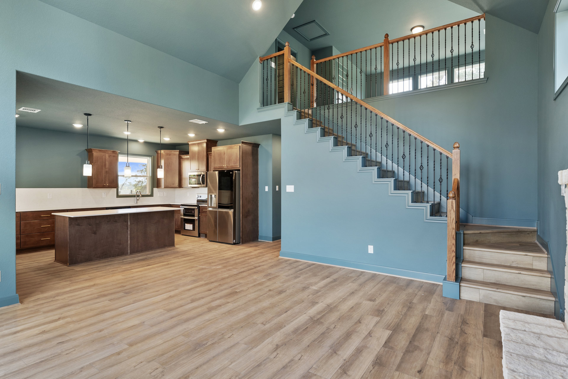 Open-concept kitchen with hardwood floors, stainless steel refrigerator, microwave above countertop, white backsplash, and adjacent wooden staircase with metal handrail.