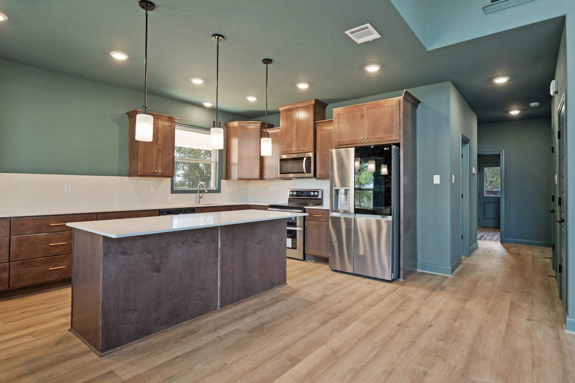 Kitchen with large white island, stainless steel refrigerator with water dispenser, white countertops, built-in microwave, white wall vent, window overlooking trees, light